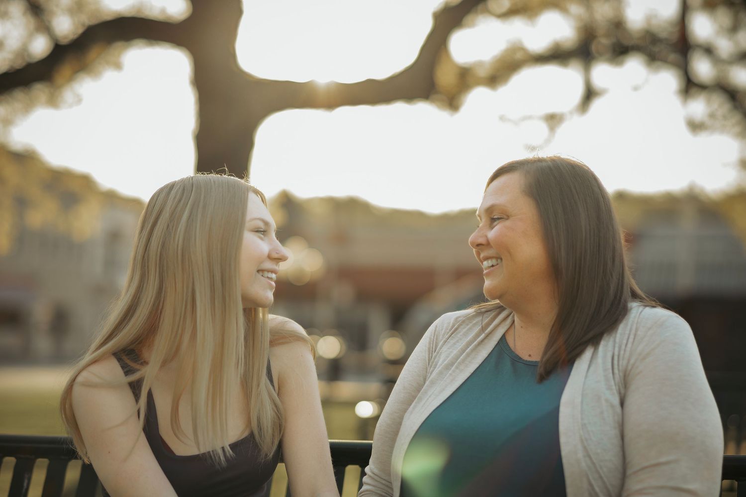 Two women smiling and conversing on a bench outdoors with warm sunset light filtering through trees behind them.