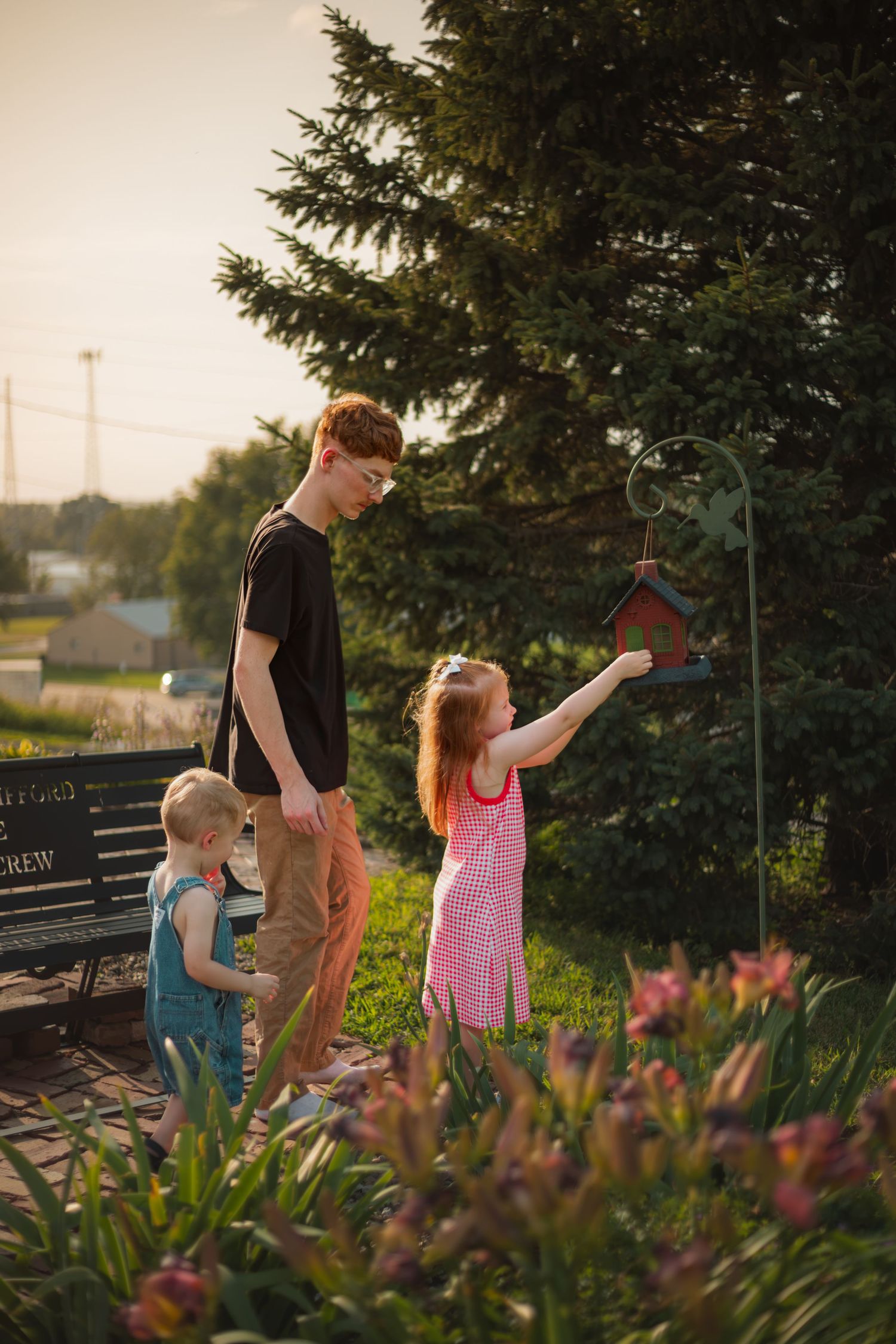 Children exploring a garden birdhouse at sunset, with a teen boy and two young girls among blooming flowers and evergreen trees.