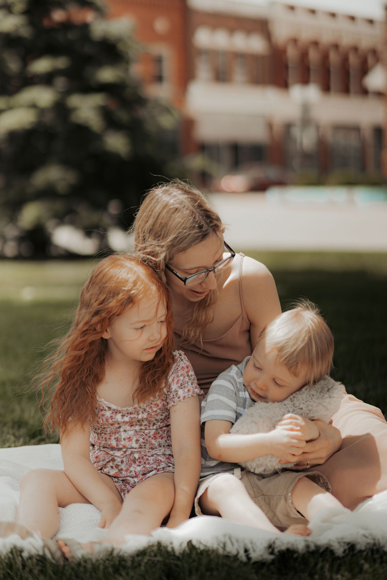 A mother with glasses sits on a blanket in a park with her red-haired daughter and young son on a sunny day.