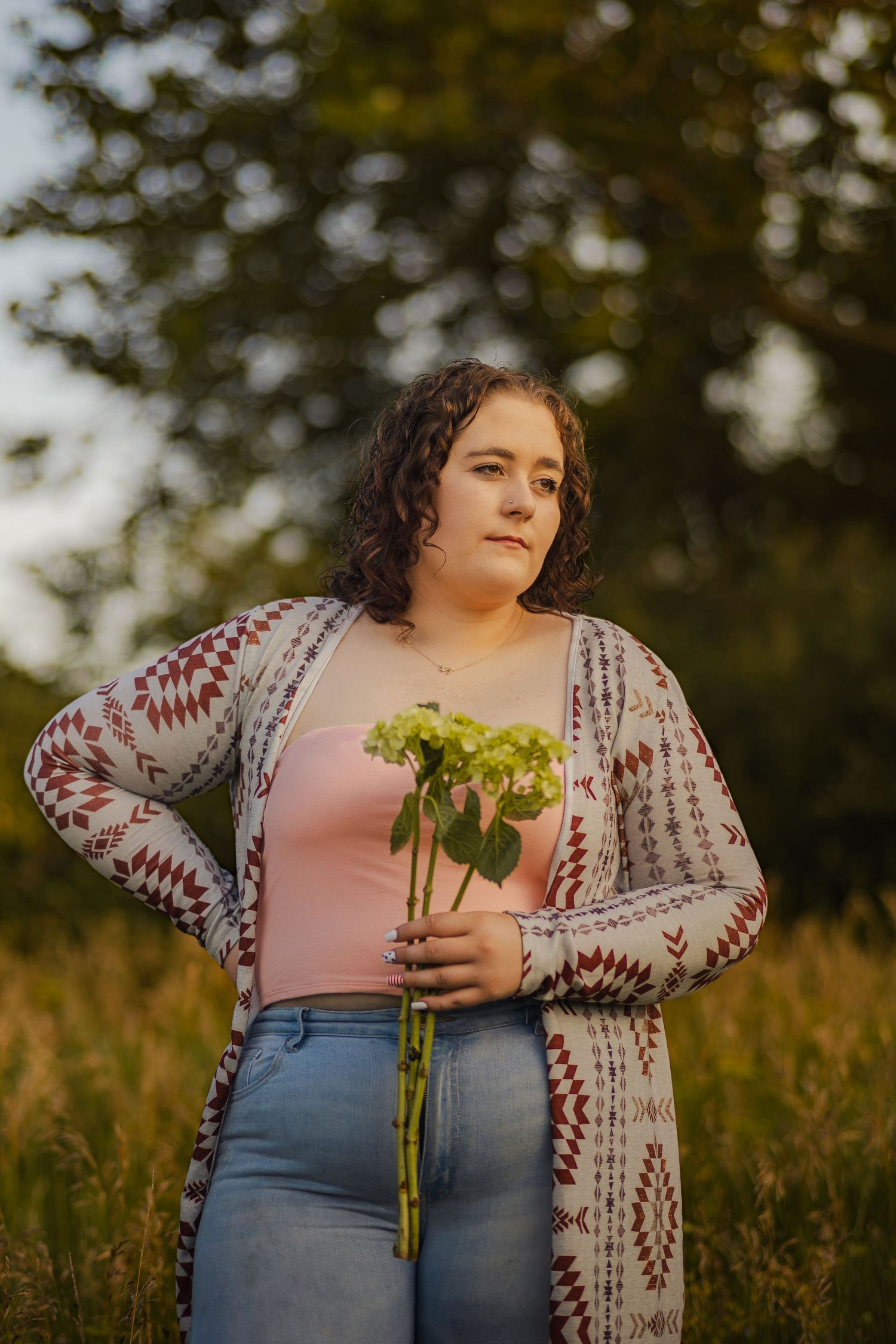 Young woman holding green flowers in a field at golden hour, wearing a patterned cardigan and jeans.