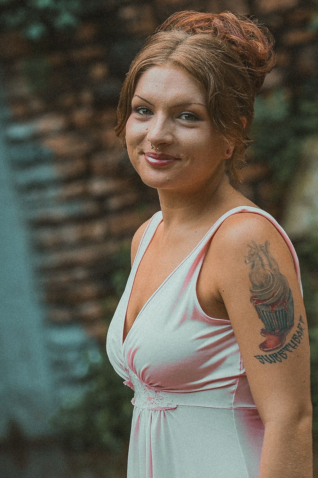 A person in a white sleeveless top poses outdoors with a visible arm tattoo against a blurred natural background.