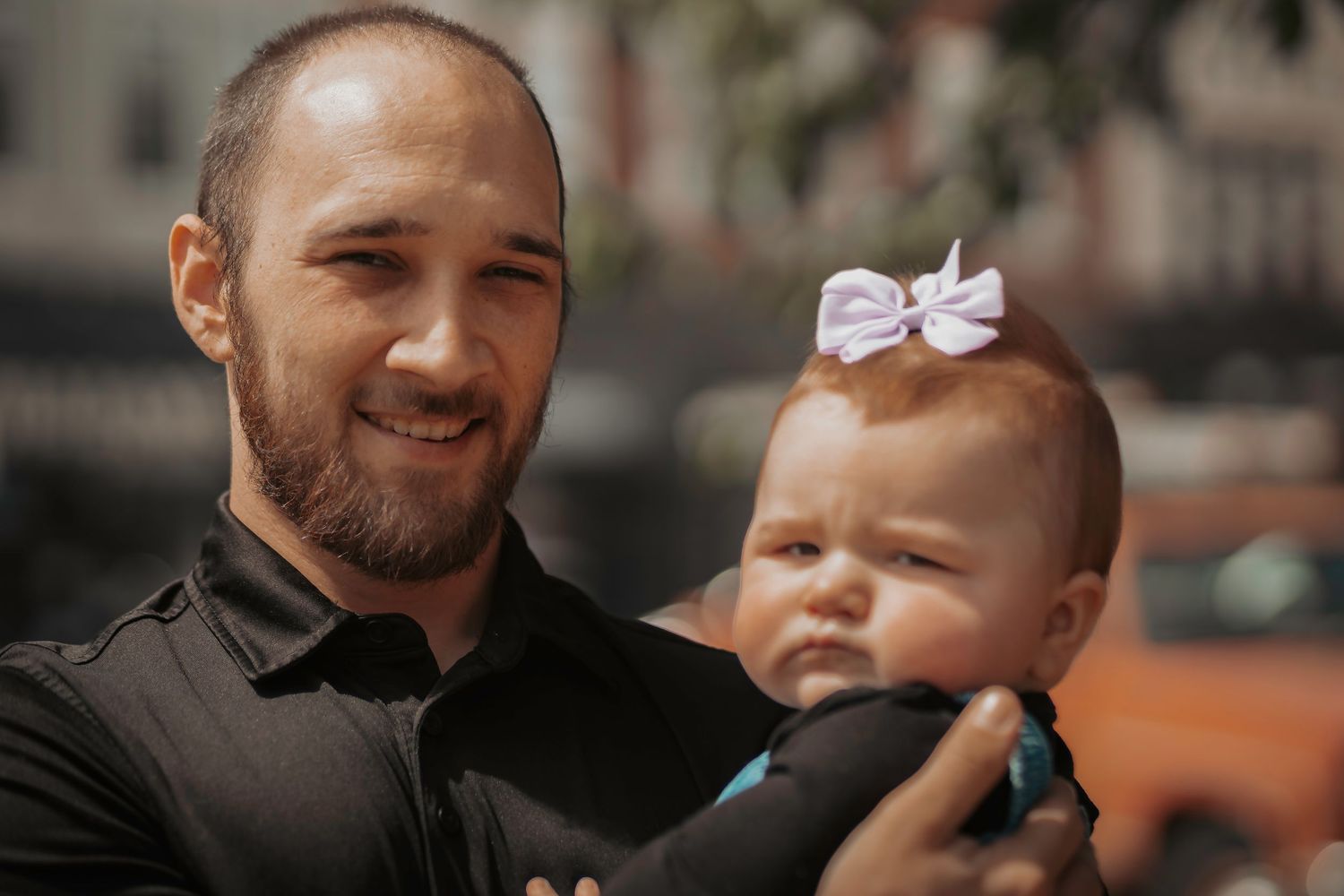 A smiling bearded man holds a baby girl wearing a white bow outdoors in a blurred urban setting.