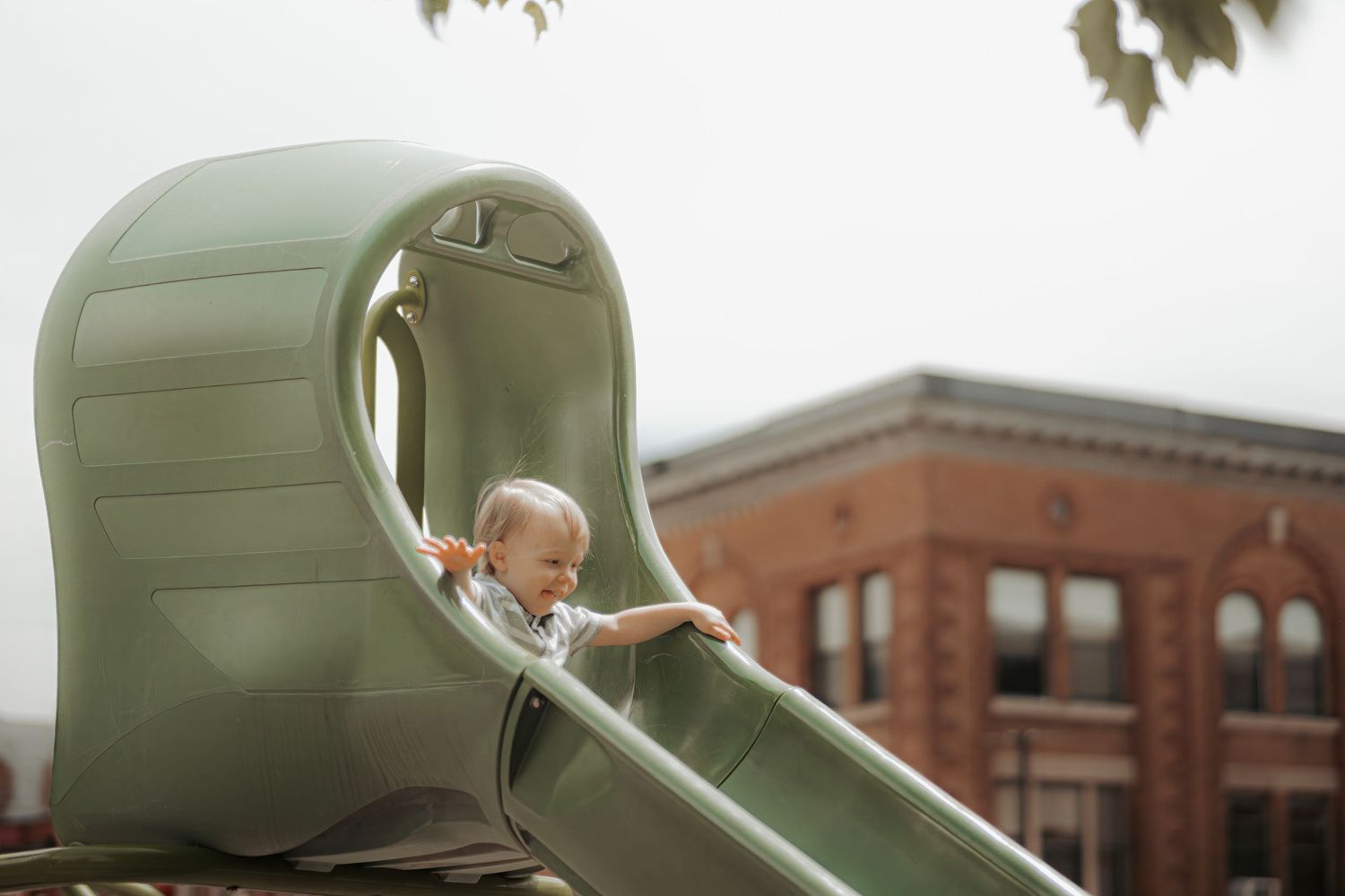 Toddler smiling while sliding down a green playground slide outdoors near a brick building.