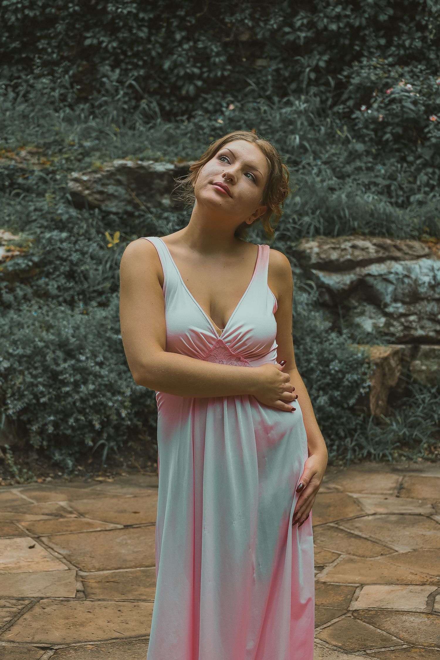 A person in a flowing pink and white ombre dress poses thoughtfully on a stone patio surrounded by greenery.