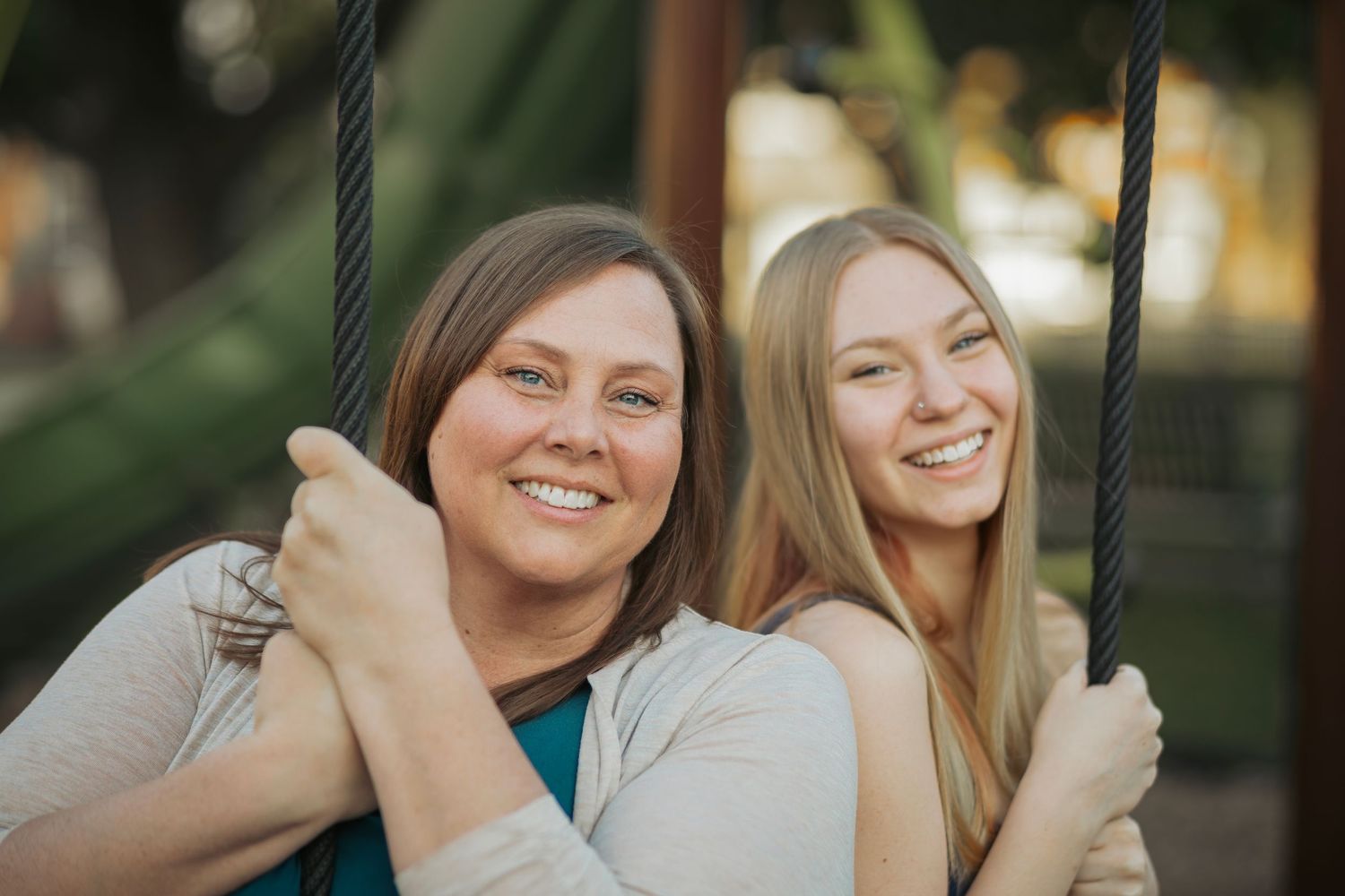 Two smiling women sitting on a swing outdoors, enjoying a sunny day together.