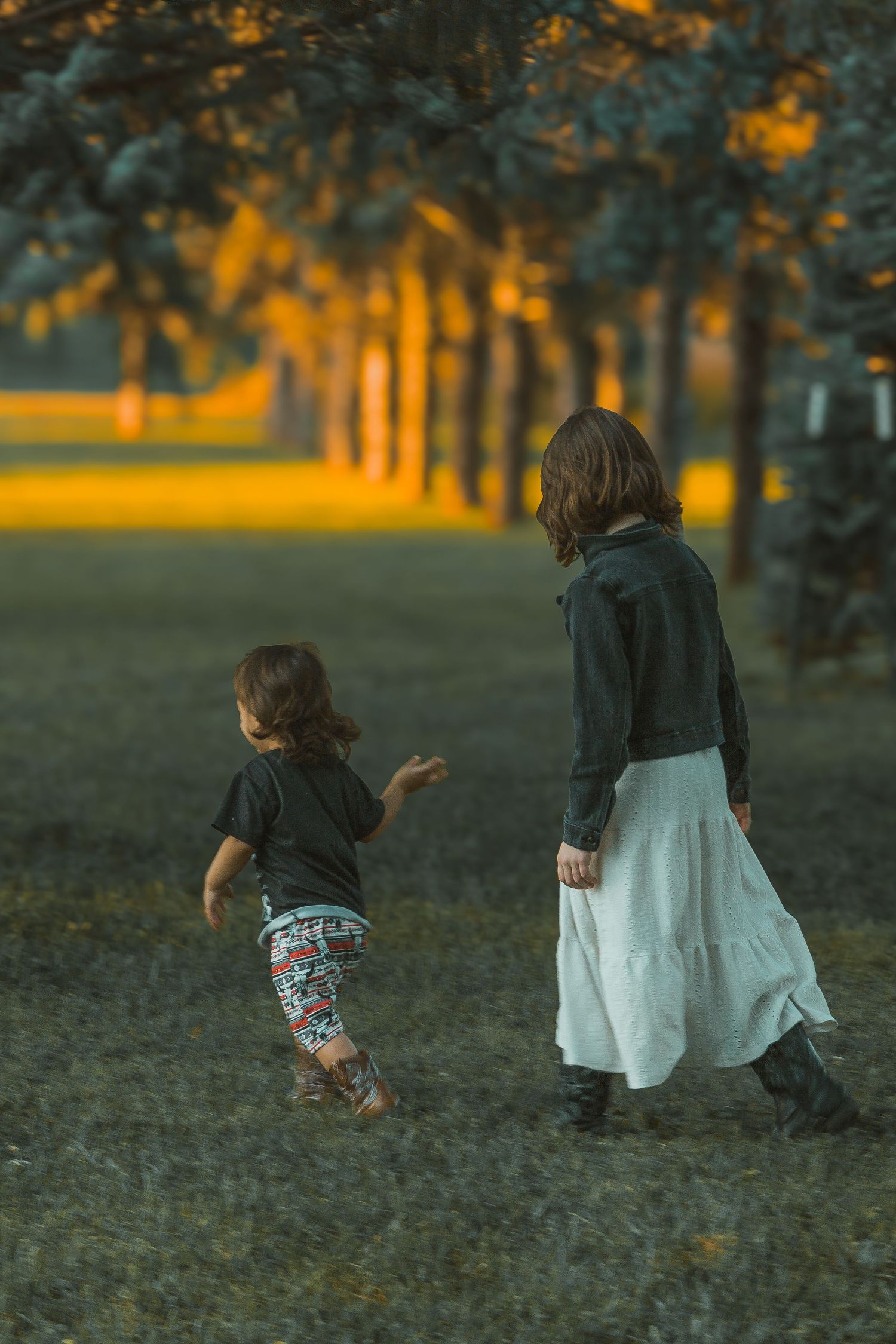 An older sibling and small toddler walk together at sunset in a park with golden light filtering through trees in the background.