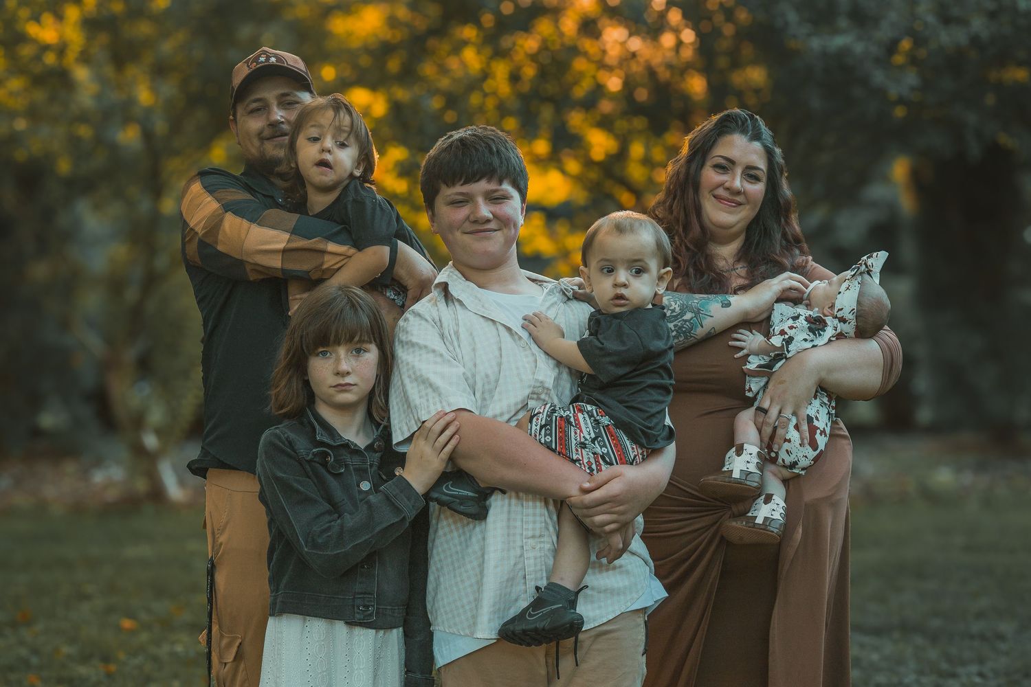 A family of six poses outdoors among autumn foliage, with parents holding young children in a warm, candid portrait.