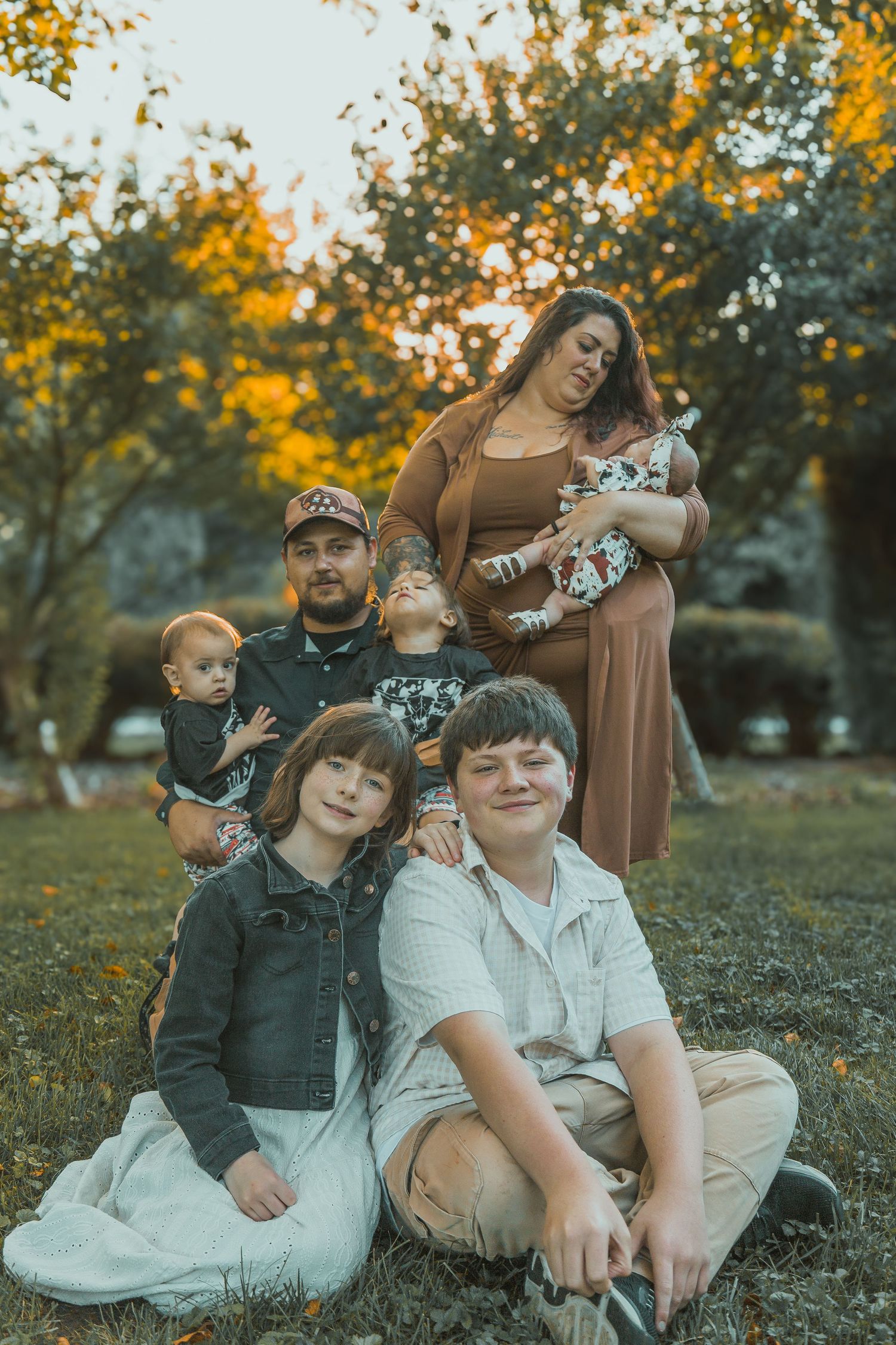 A family sits together outdoors during the golden hour of an autumn sunset, with fall foliage in the background.