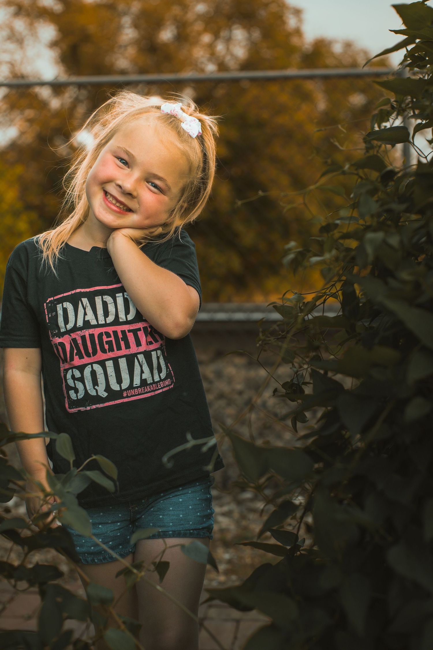Smiling blonde girl in a black Daddy Daughter Squad t-shirt posing outdoors among green foliage in warm autumn light.