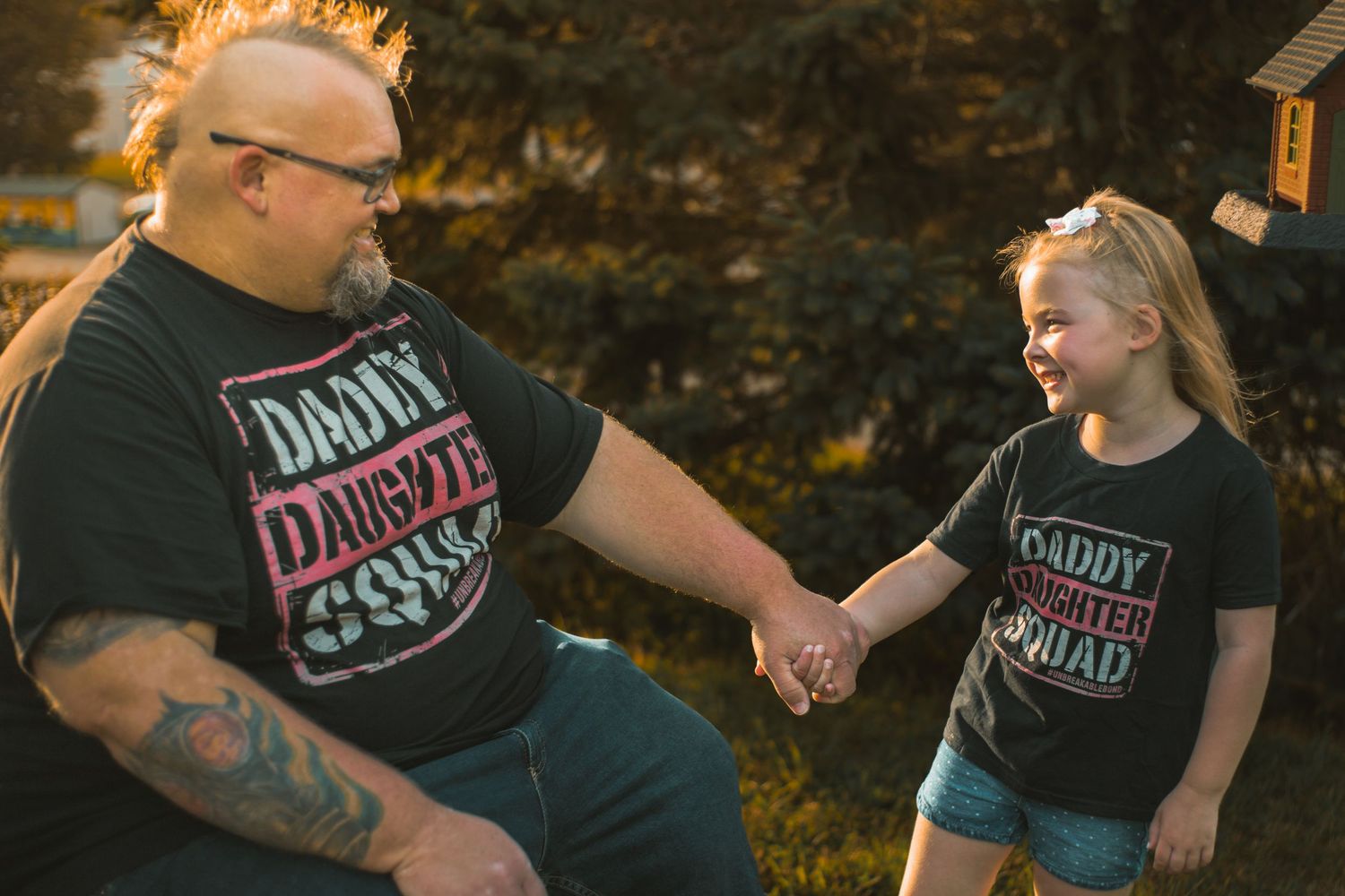 Father and daughter in matching Daddy Daughter Squad black t-shirts holding hands and smiling outdoors.