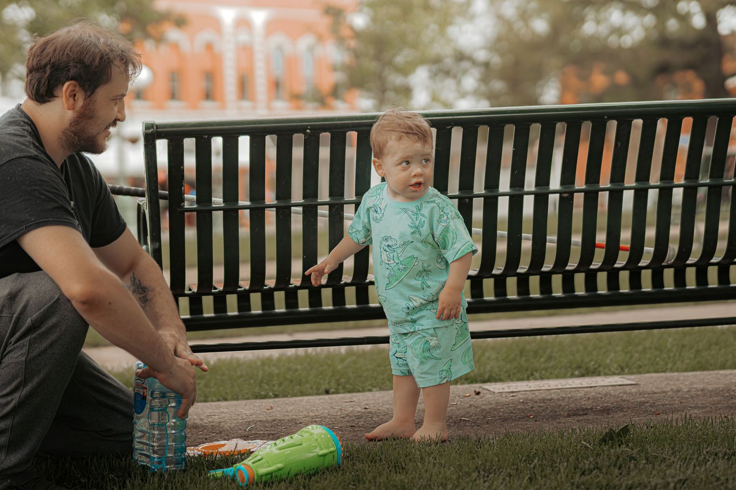A father crouches beside his toddler in a green outfit near a park bench on a grassy lawn.