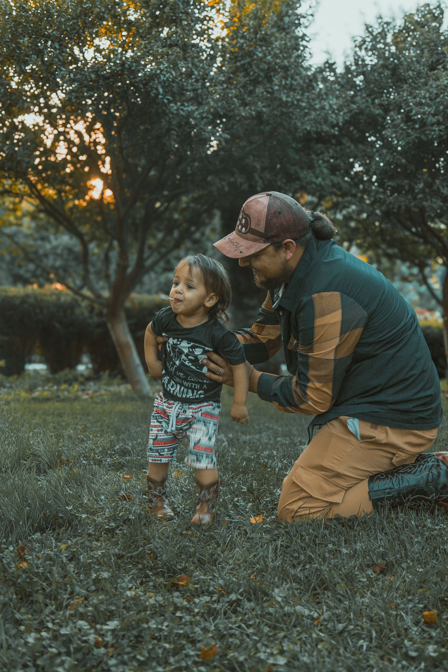A father in a brown jacket crouches low to capture his rambunctious small child in a grassy field at sunset.