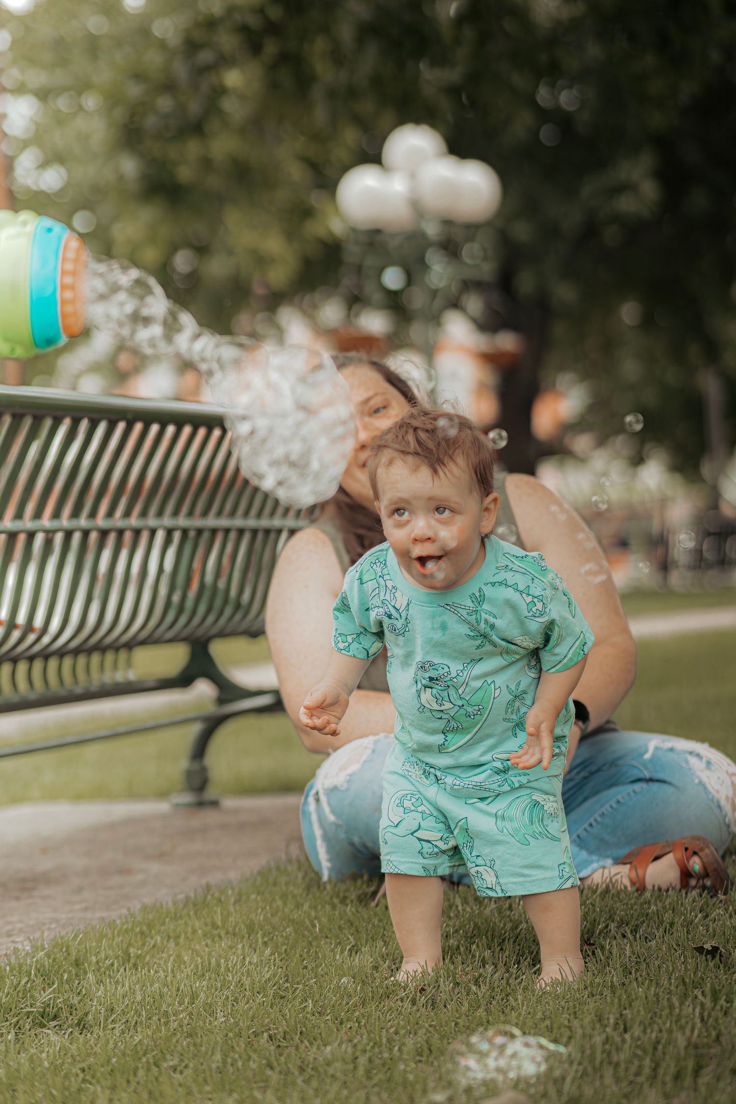 A mother kisses her toddler boy outdoors on grass near a park bench while bubbles float around them.