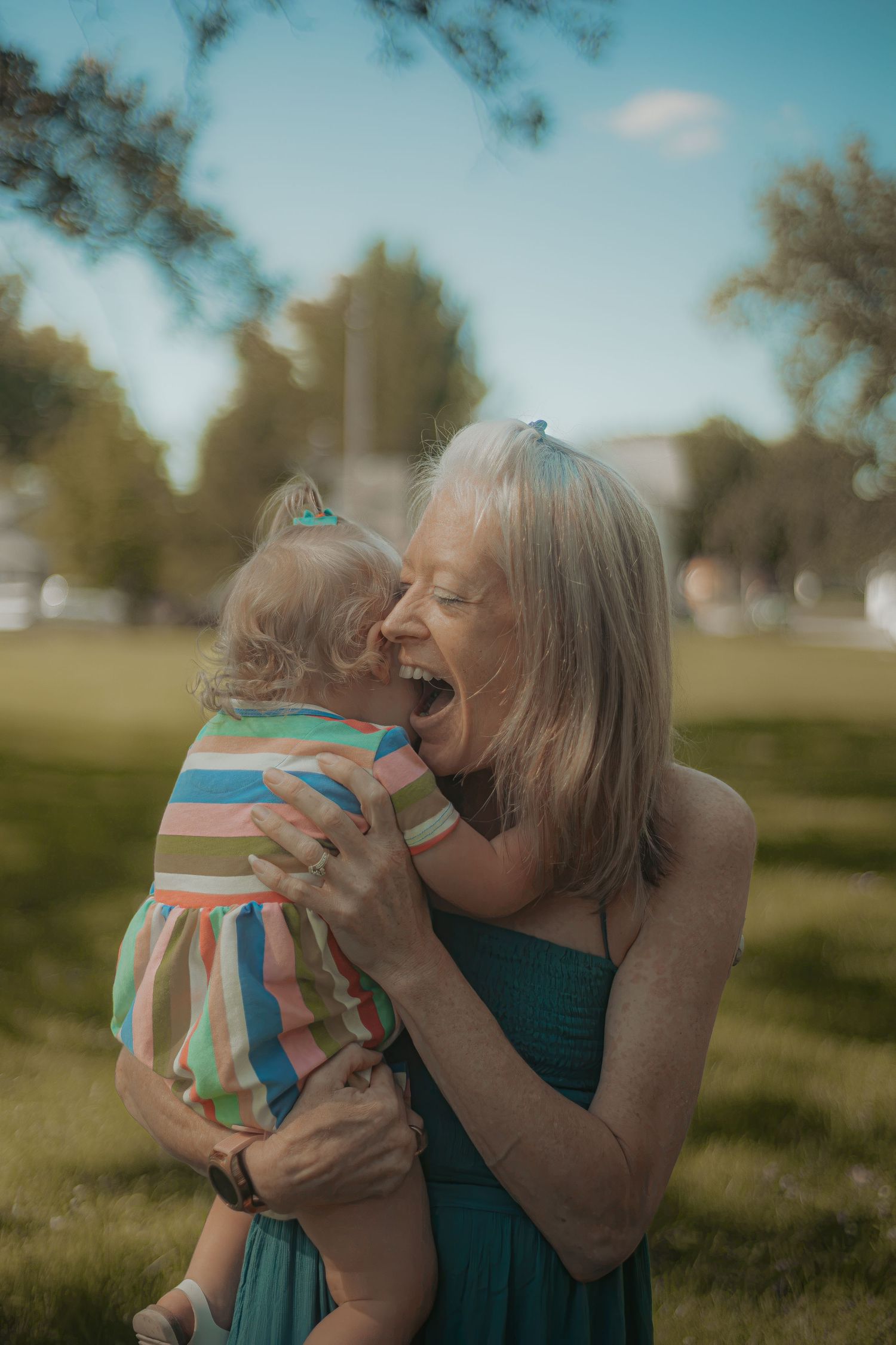 Grandmother laughing joyfully while holding a baby in a colorful striped dress outdoors in a sunny park.