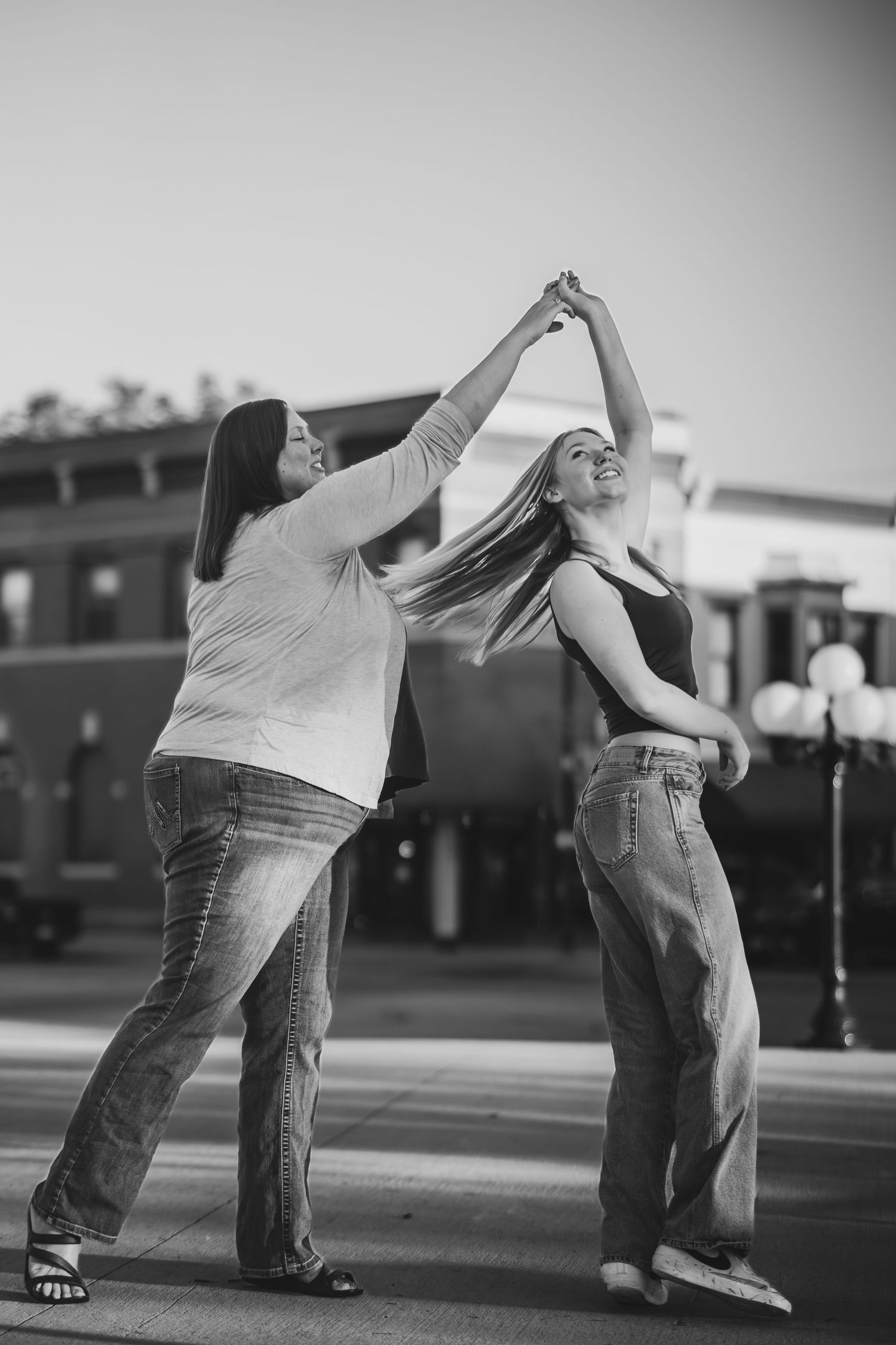 Two women dancing outdoors in black and white photo, one spinning the other near a train station.