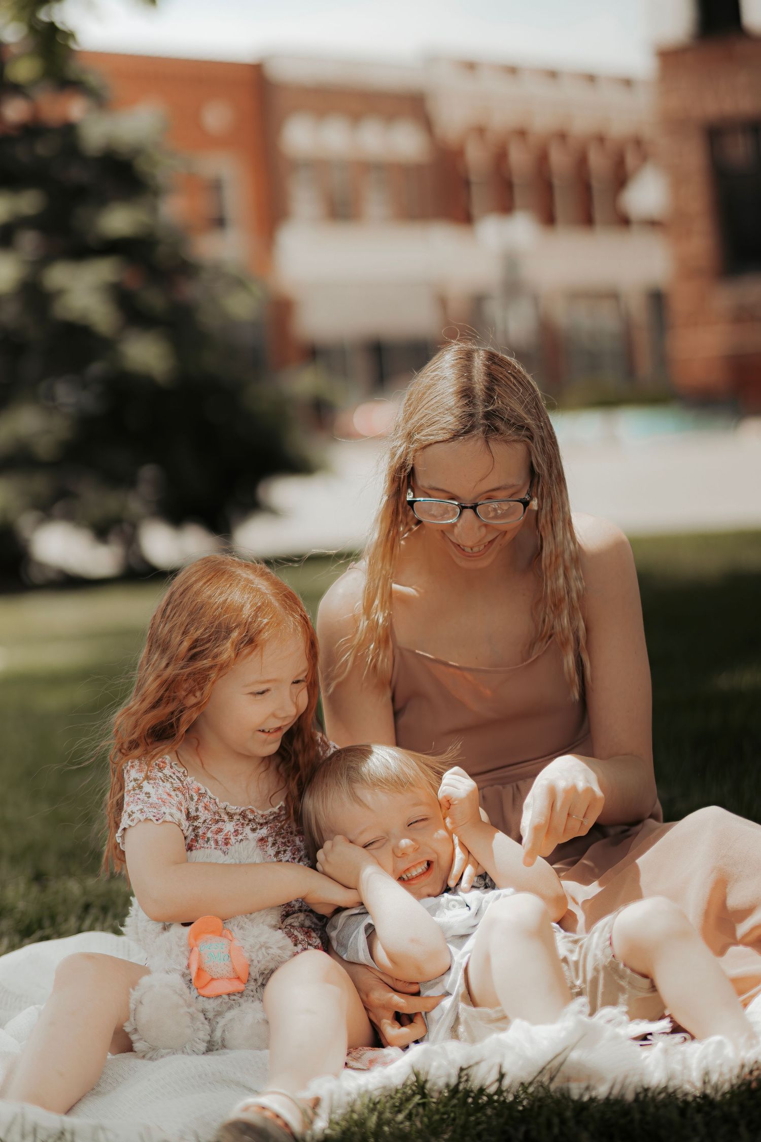 A mother and two young children sitting on a blanket outdoors on a sunny day, enjoying time together in a park.