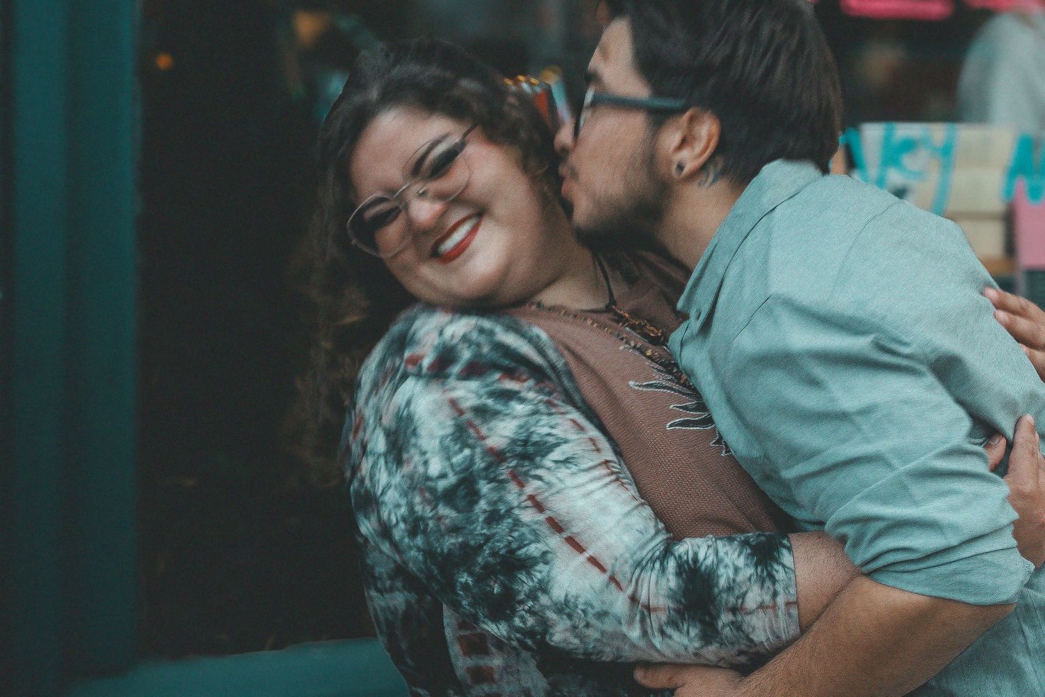 A happy couple sharing a tender moment, man kissing woman on cheek while she smiles joyfully outdoors.