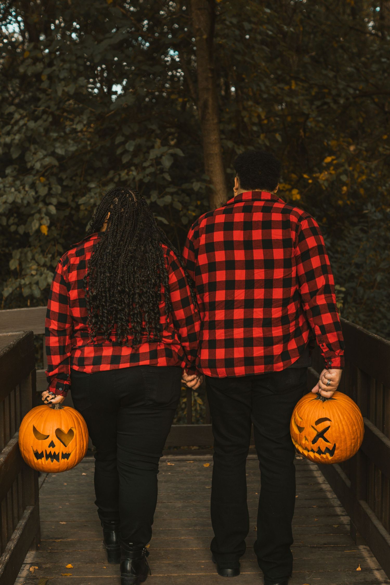 Couple in matching red plaid shirts holding carved Halloween jack-o-lanterns while walking on a wooden bridge.