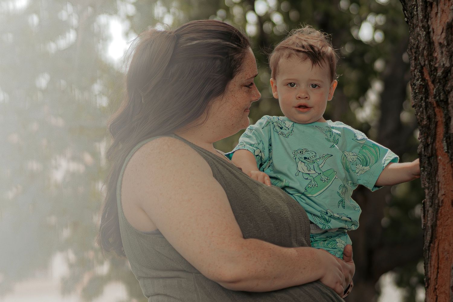 A mother lovingly holds her baby boy in a teal outfit near a tree in a soft, misty outdoor setting.