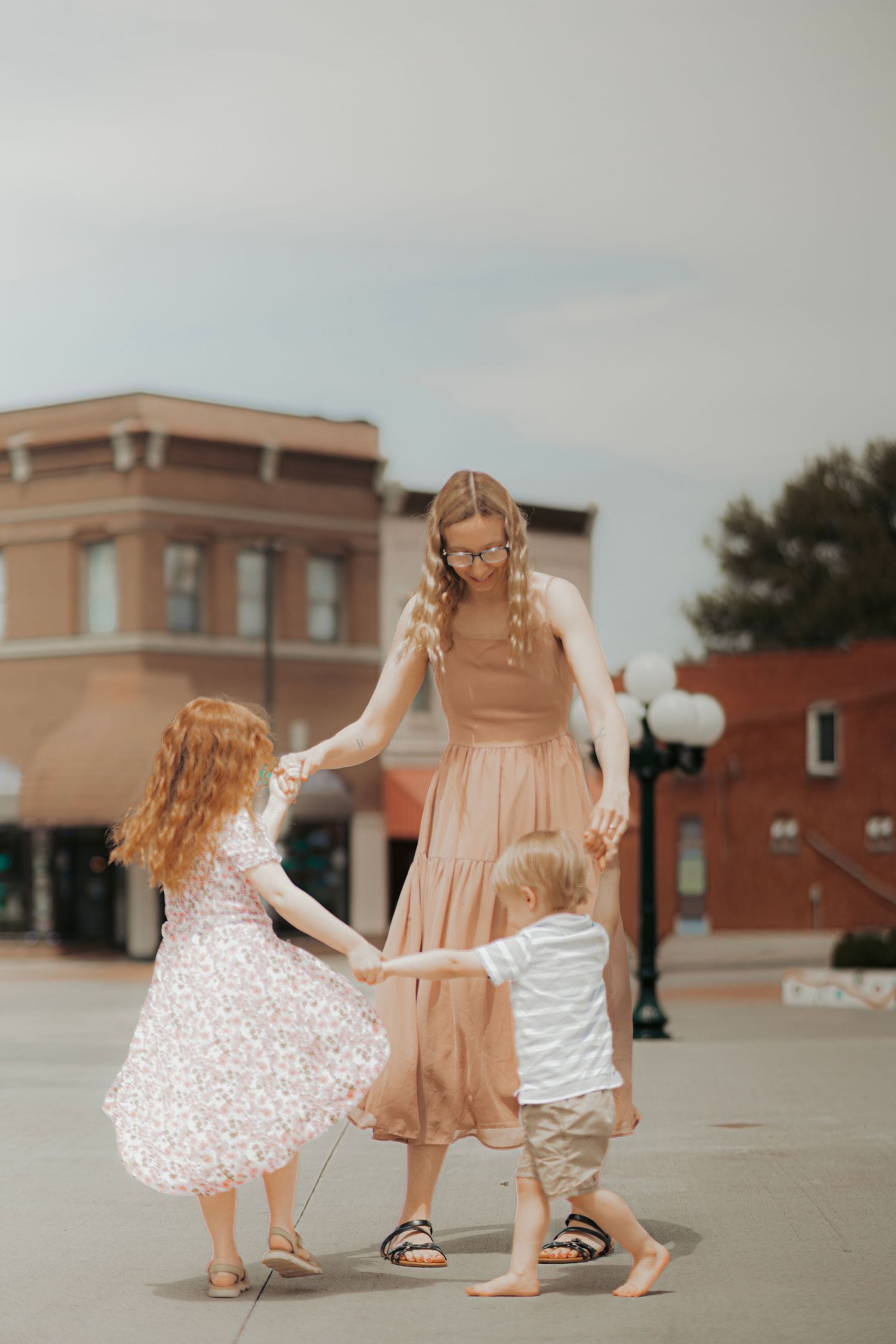 A mother in a tan dress dances with her two young children on a town square sidewalk on a cloudy day.