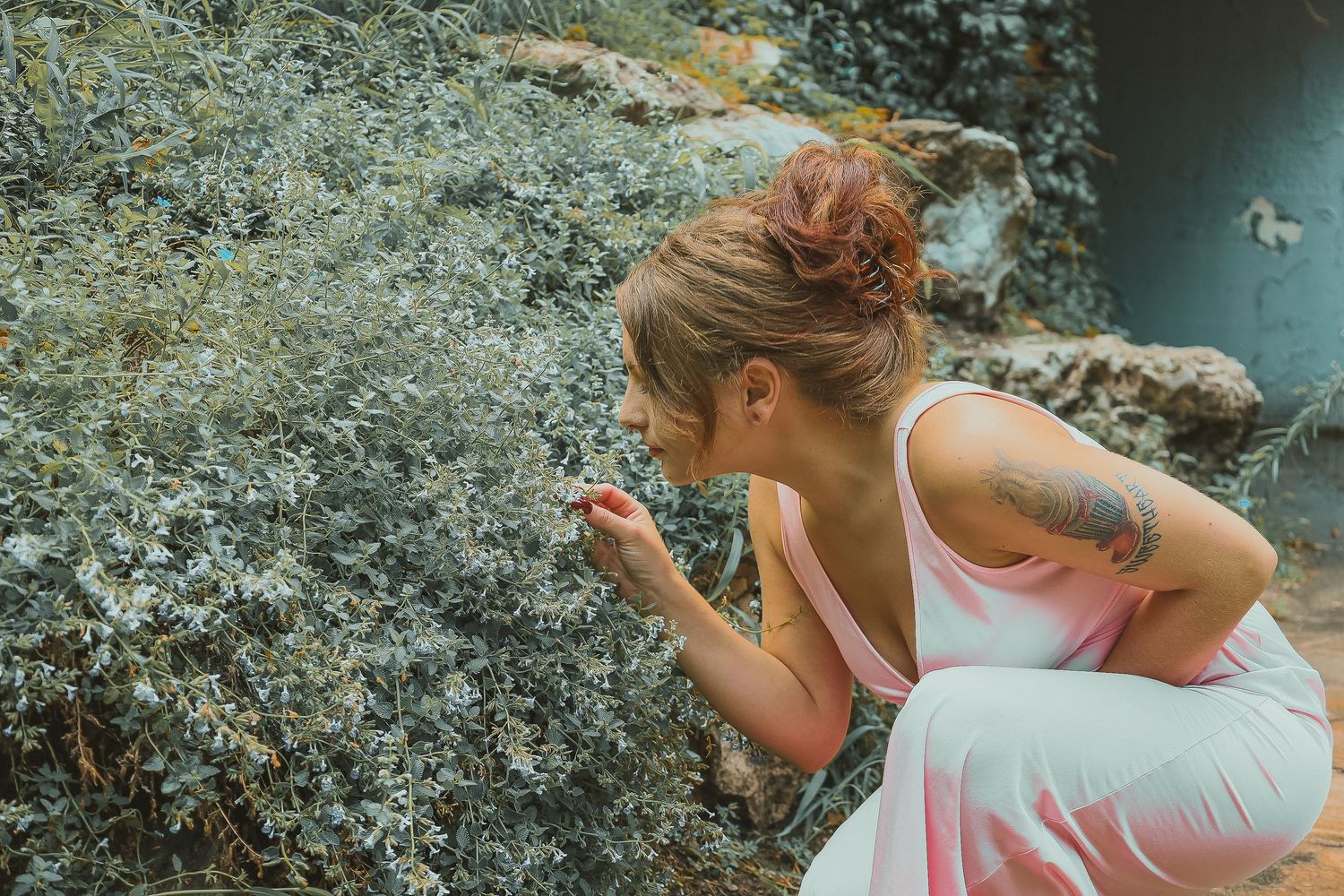 Someone in a white tank top and an upper arm tattoo leans down to smell wildflowers growing among rocks in nature.