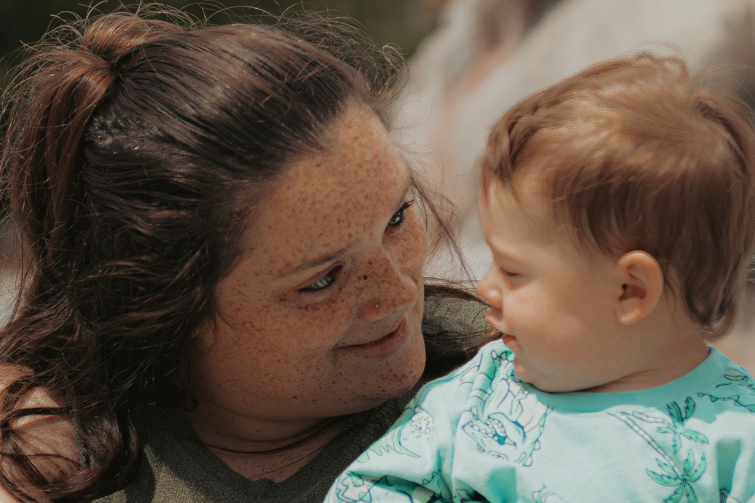 A freckled brunette woman tenderly gazes at a smiling baby in a teal outfit.