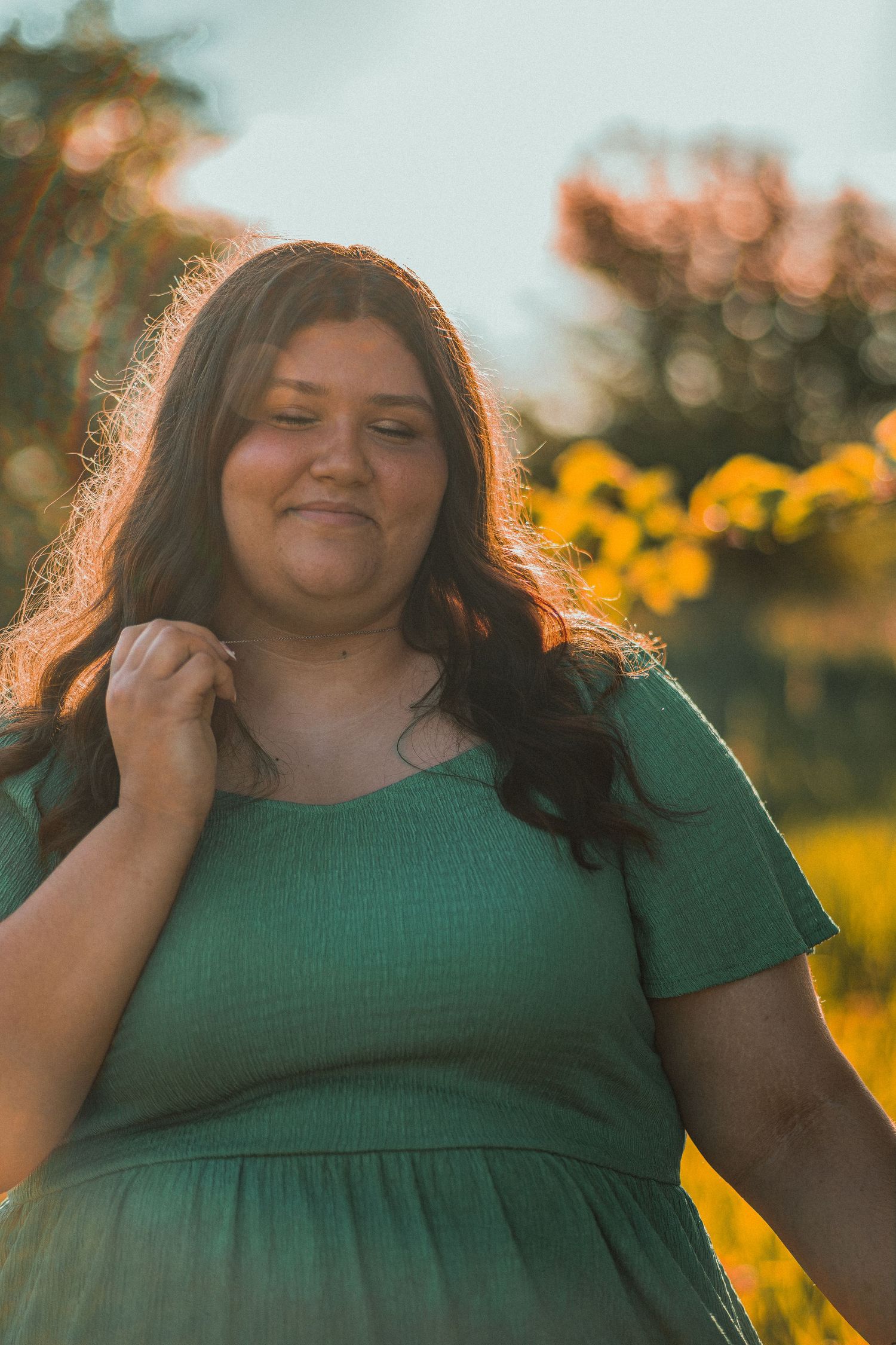 Young woman in green dress smiling outdoors at golden hour with bokeh sunflowers in background.