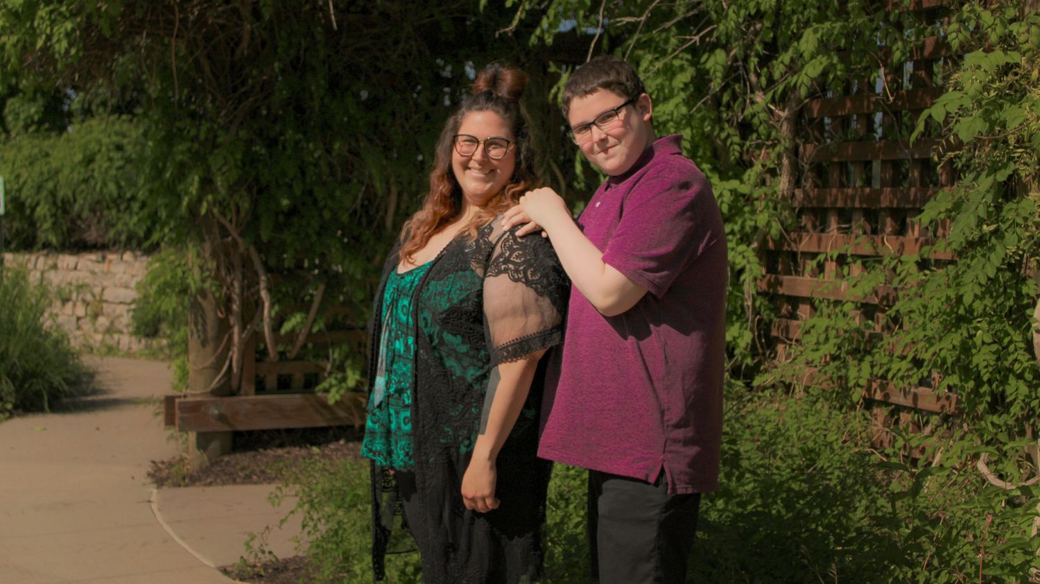 A mother and son pose together on a garden path during a sunny outdoor photoshoot.