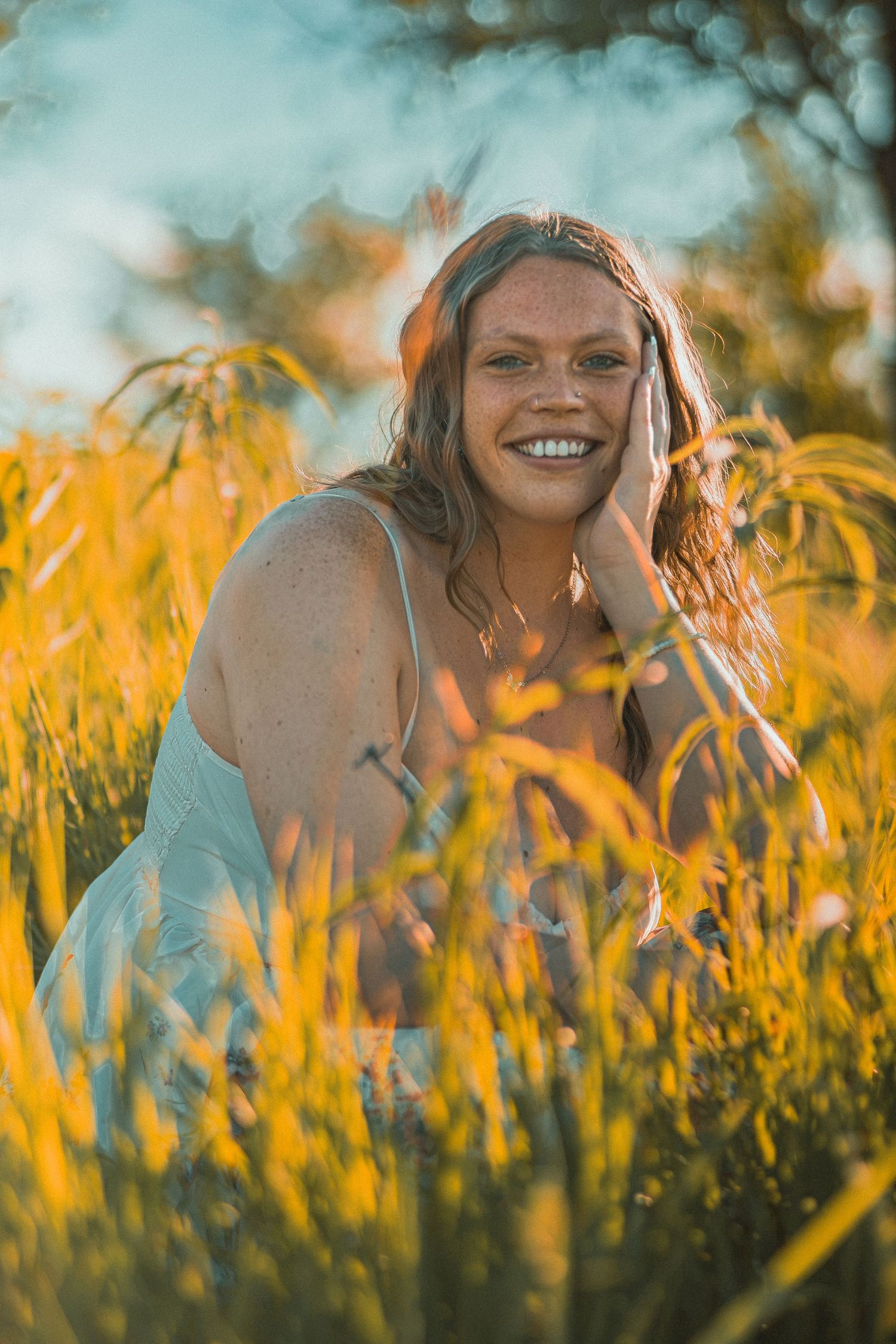 A smiling woman with red hair sits in golden tall grass during sunset, hand on cheek, wearing a white dress.