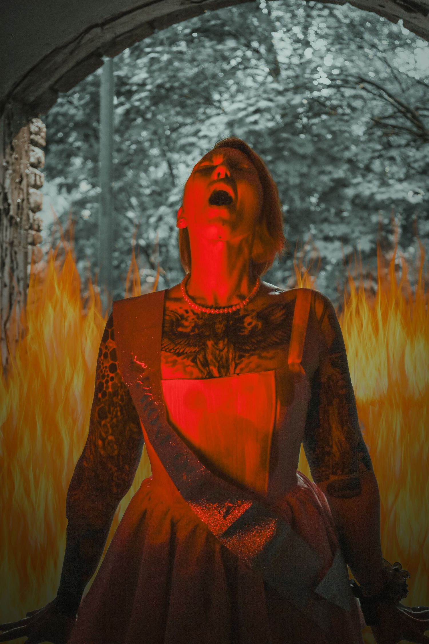 Tattooed woman in red dress with mouth open, surrounded by flames in dramatic dark artistic photo.