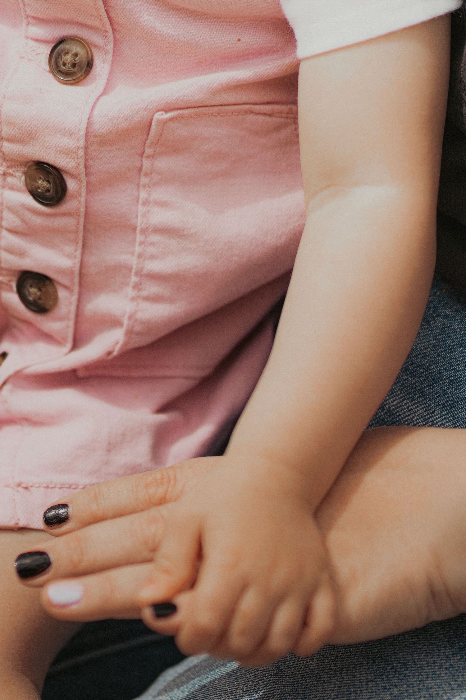 Child in pink button-up romper sits beside adult with dark nail polish, hands gently touching.