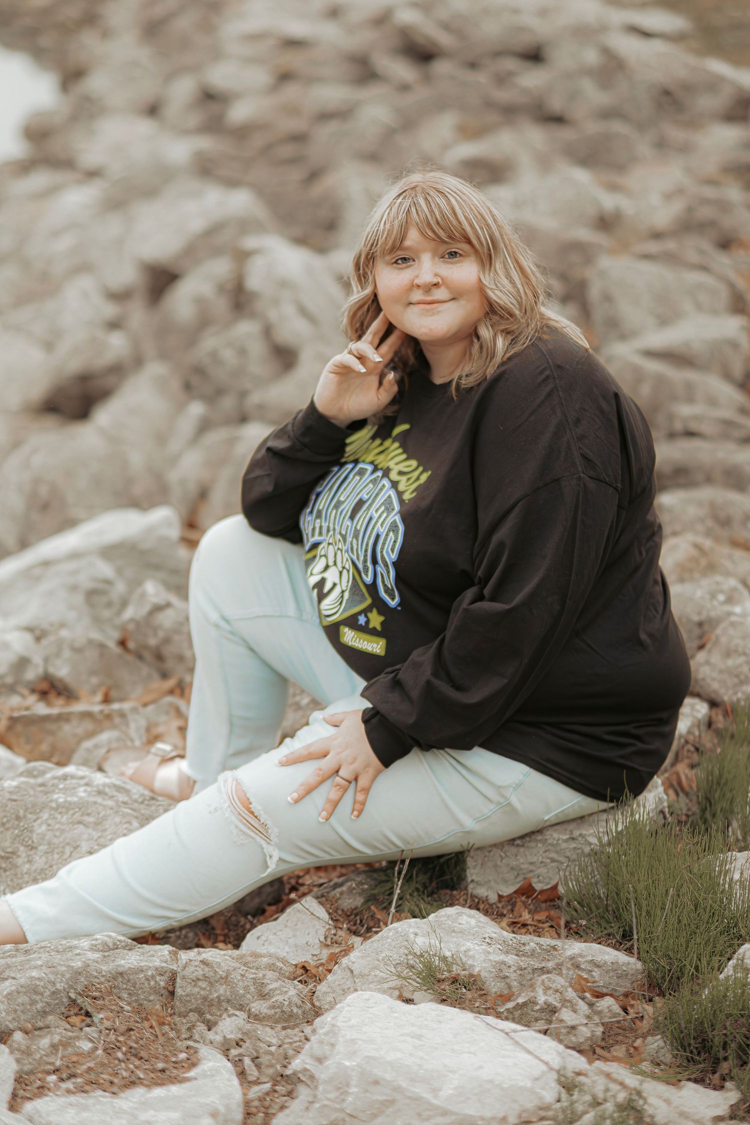 Young woman in black graphic sweatshirt and white ripped jeans sitting on rocks making a peace sign.