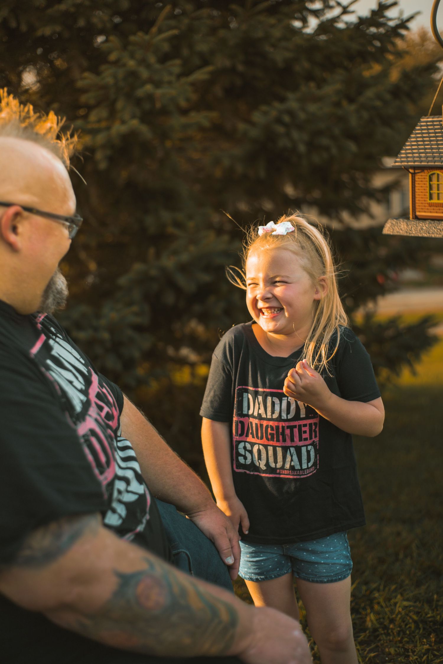 A young girl in a Daddy Daughter Squad shirt laughs joyfully beside her tattooed father outdoors at golden hour.