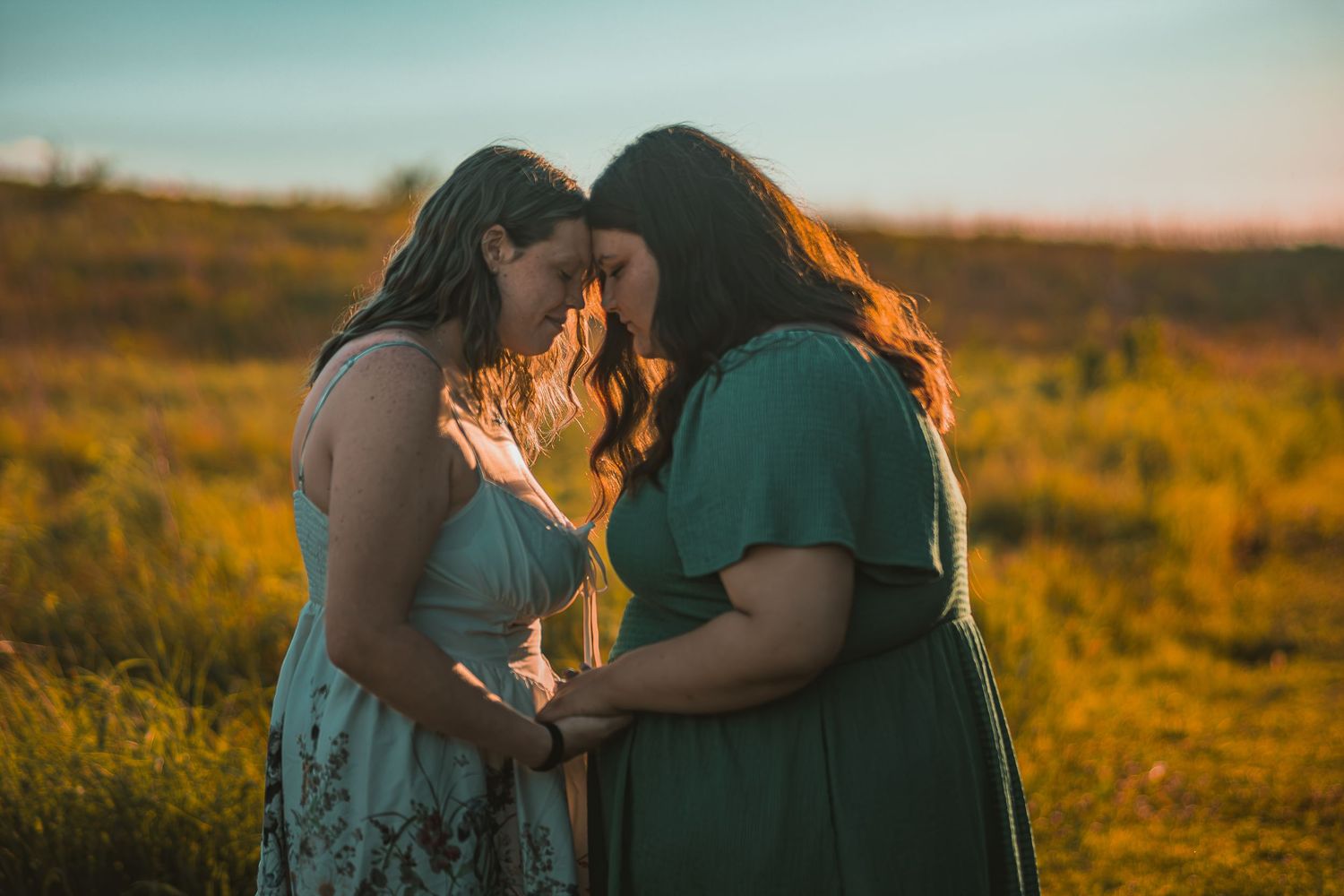 Two women in floral and green dresses touch foreheads in a golden field at sunset.