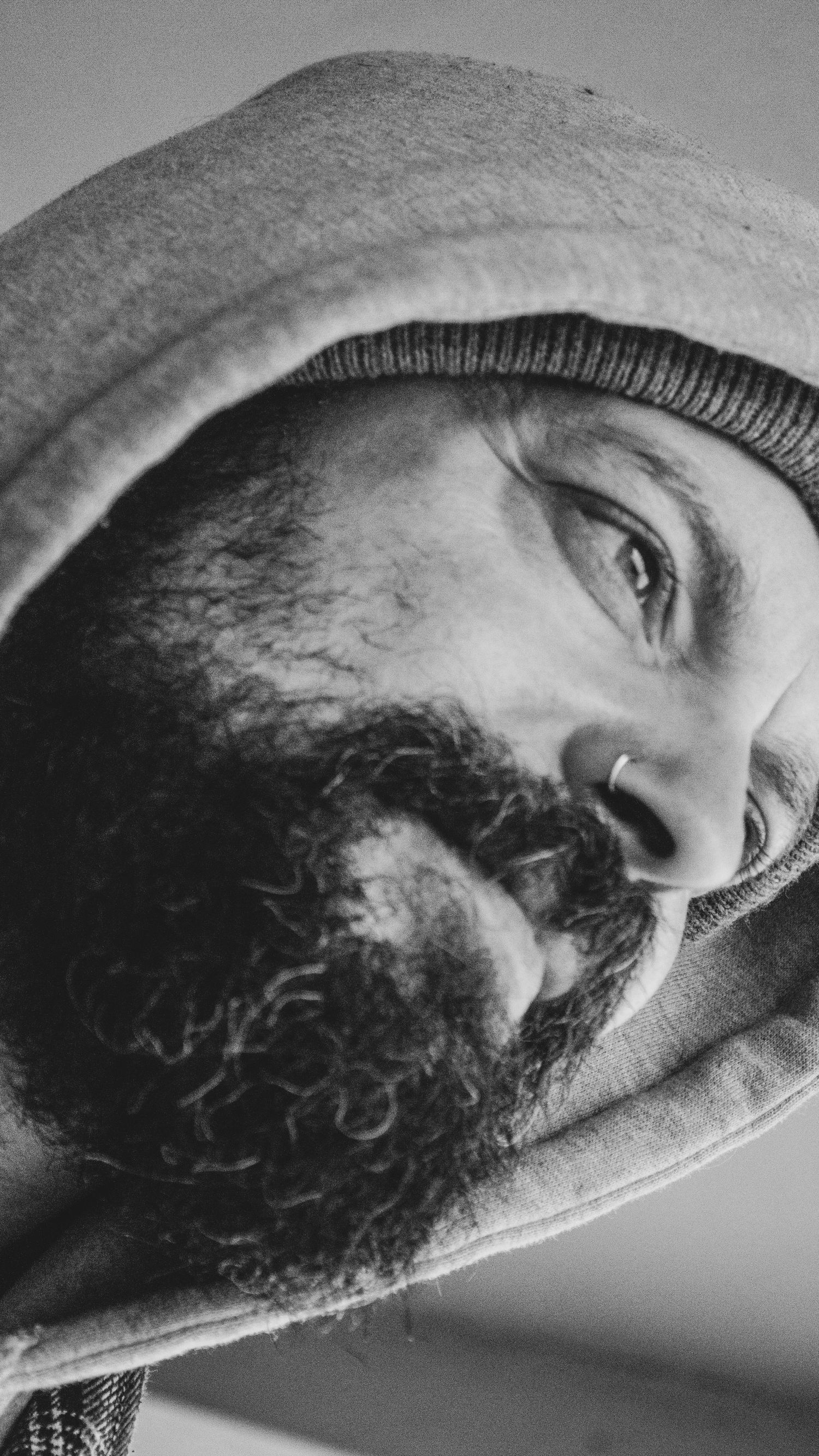 Close-up black and white portrait of a bearded person wearing a knit beanie, looking at the camera from an angle.