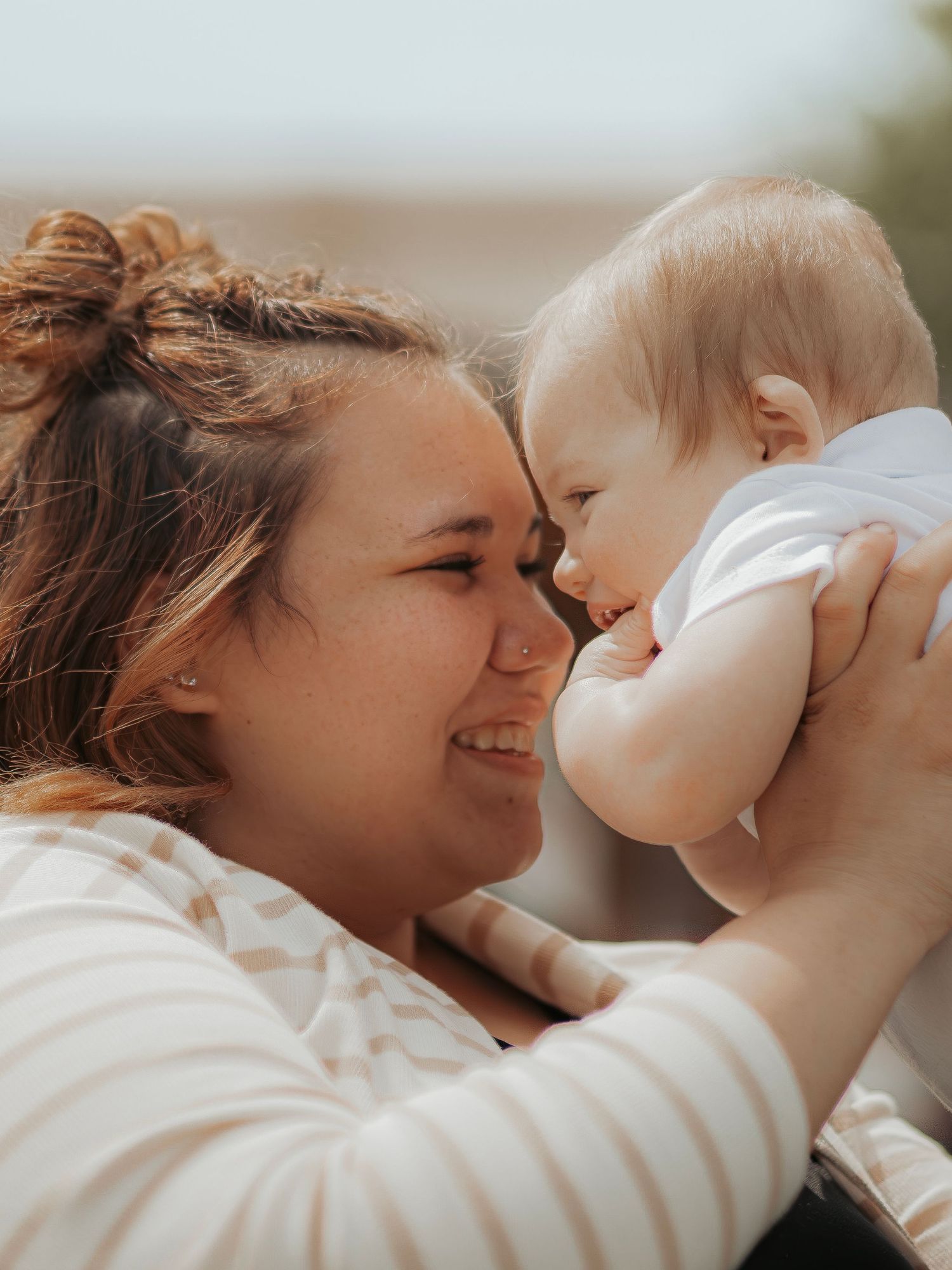 A smiling young woman holds a happy baby close to her face in a warm, sunny outdoor setting.