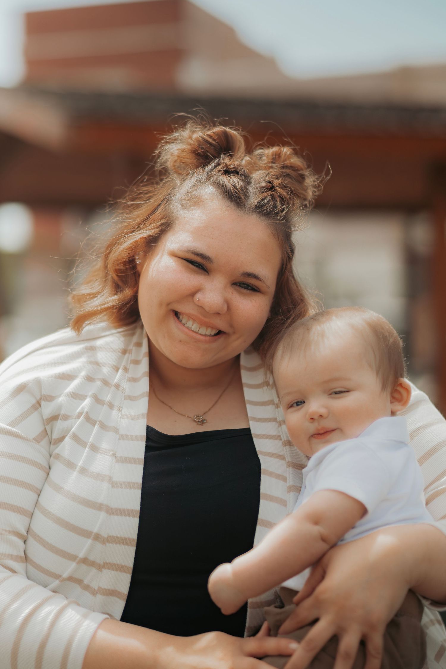 Smiling young woman with bun hairstyle holding an adorable baby outdoors in natural light.