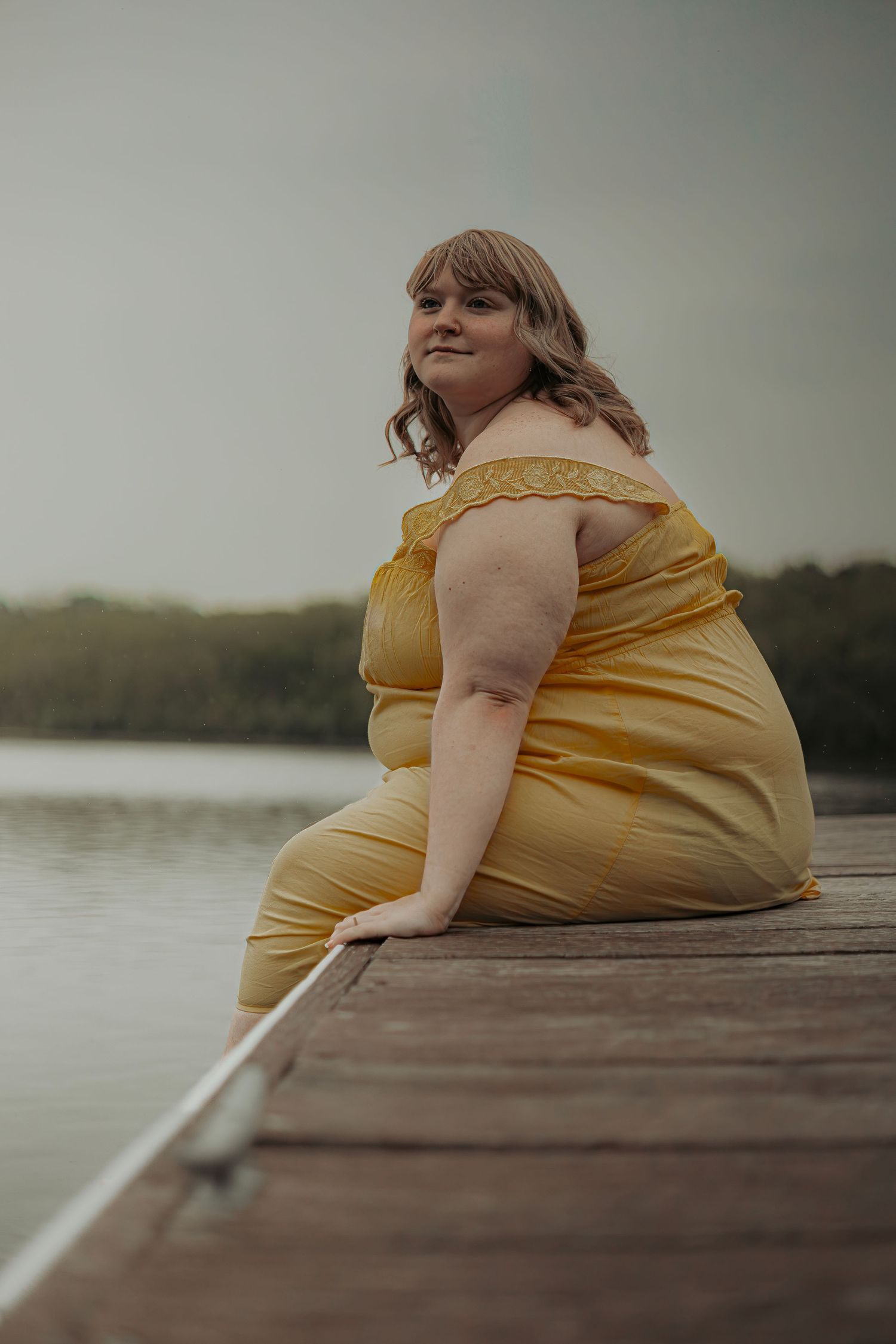 Plus-size woman in yellow off-shoulder jumpsuit sitting on wooden dock by calm lake, smiling at camera.