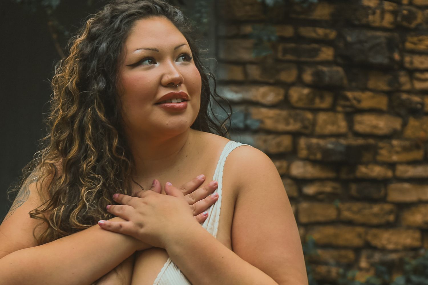 A person in a white top poses with hands crossed over the heart against a rustic brick wall background.