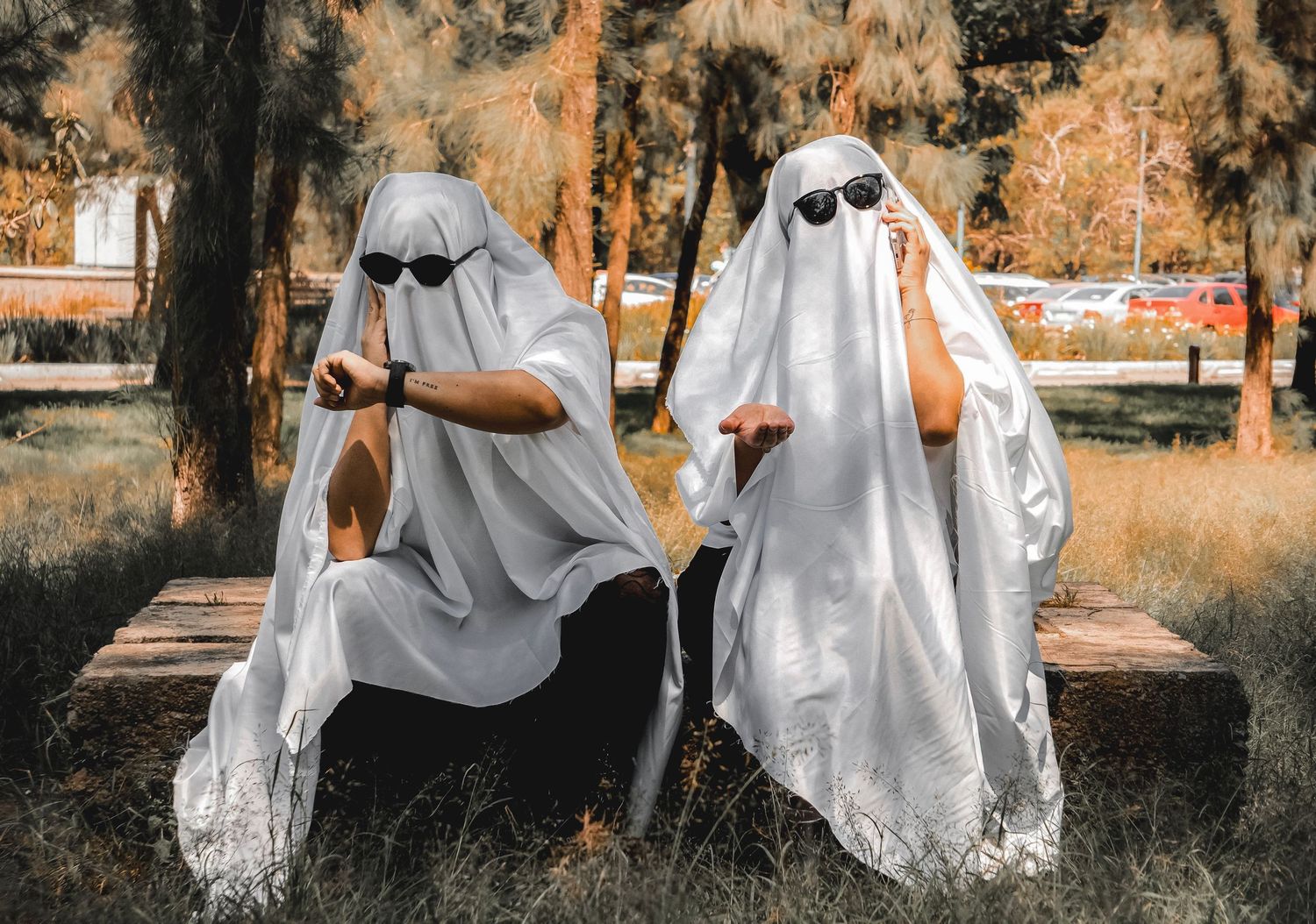 Two people dressed as ghosts in white sheets with sunglasses sitting on a bench outdoors for Halloween.