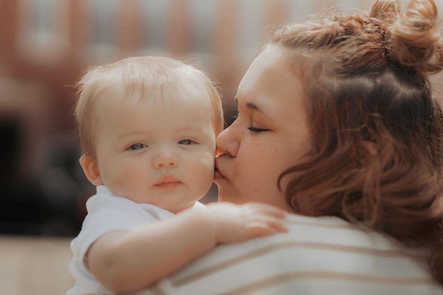 A loving mother tenderly kisses her adorable baby on the cheek in a warm, candid outdoor portrait.