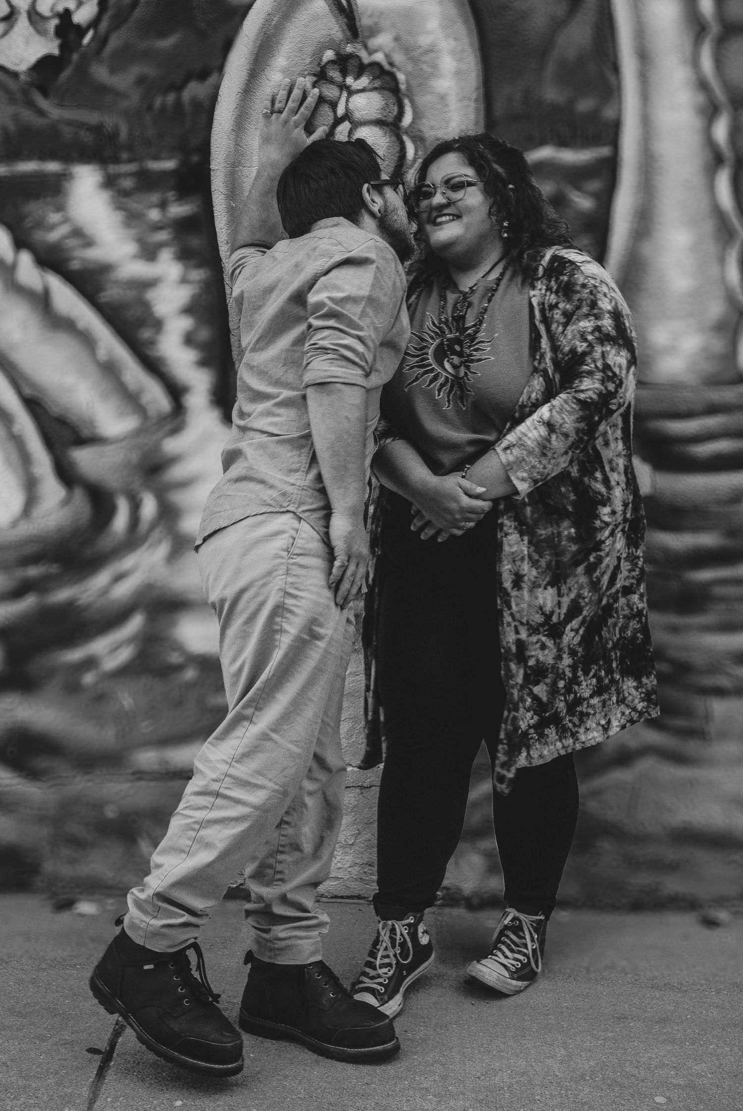 Black and white photo of a couple sharing a tender moment in front of a colorful mural outdoors.