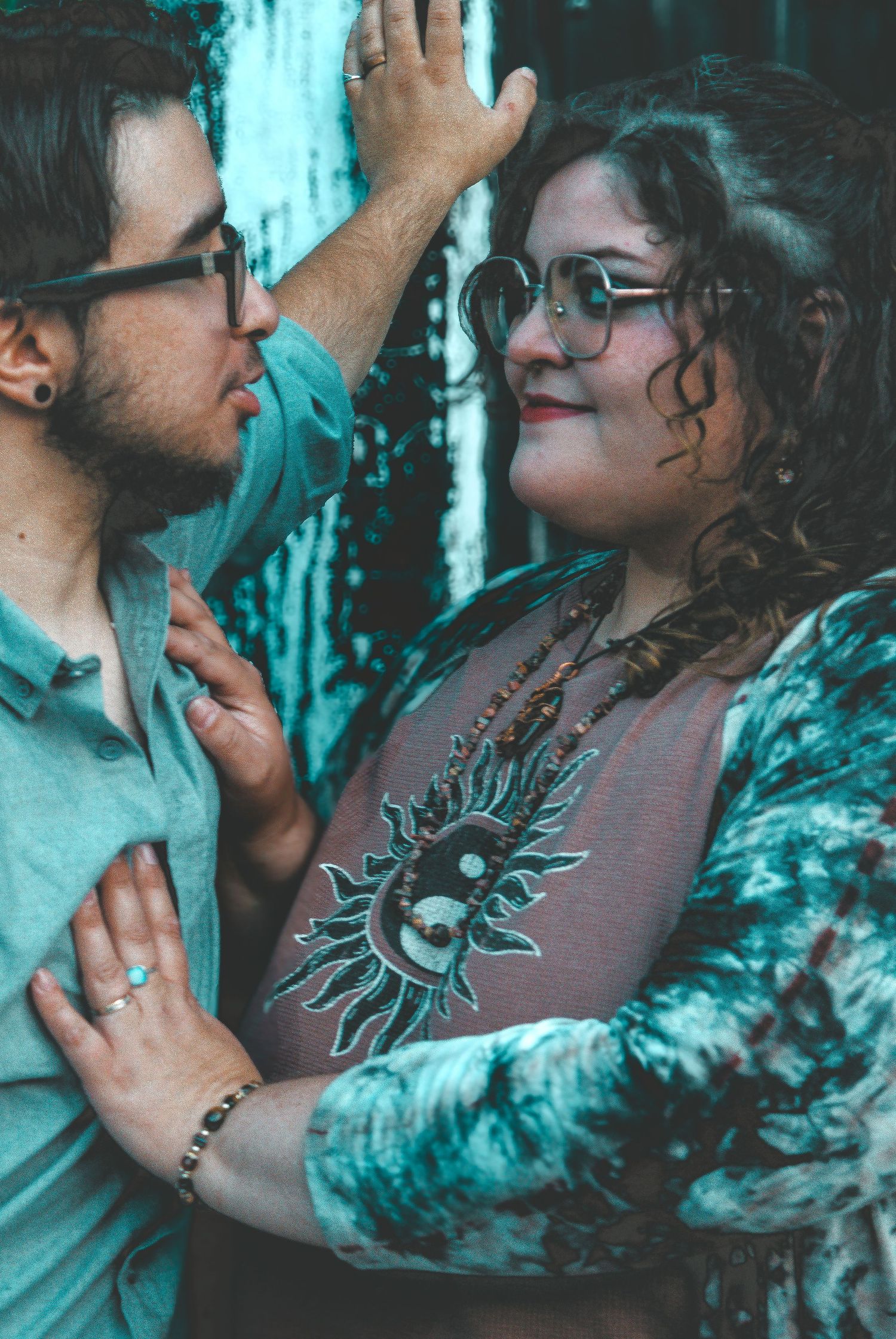 A couple in glasses share a tender moment, the man touching the woman's hair while she smiles, wearing a sun graphic tee.