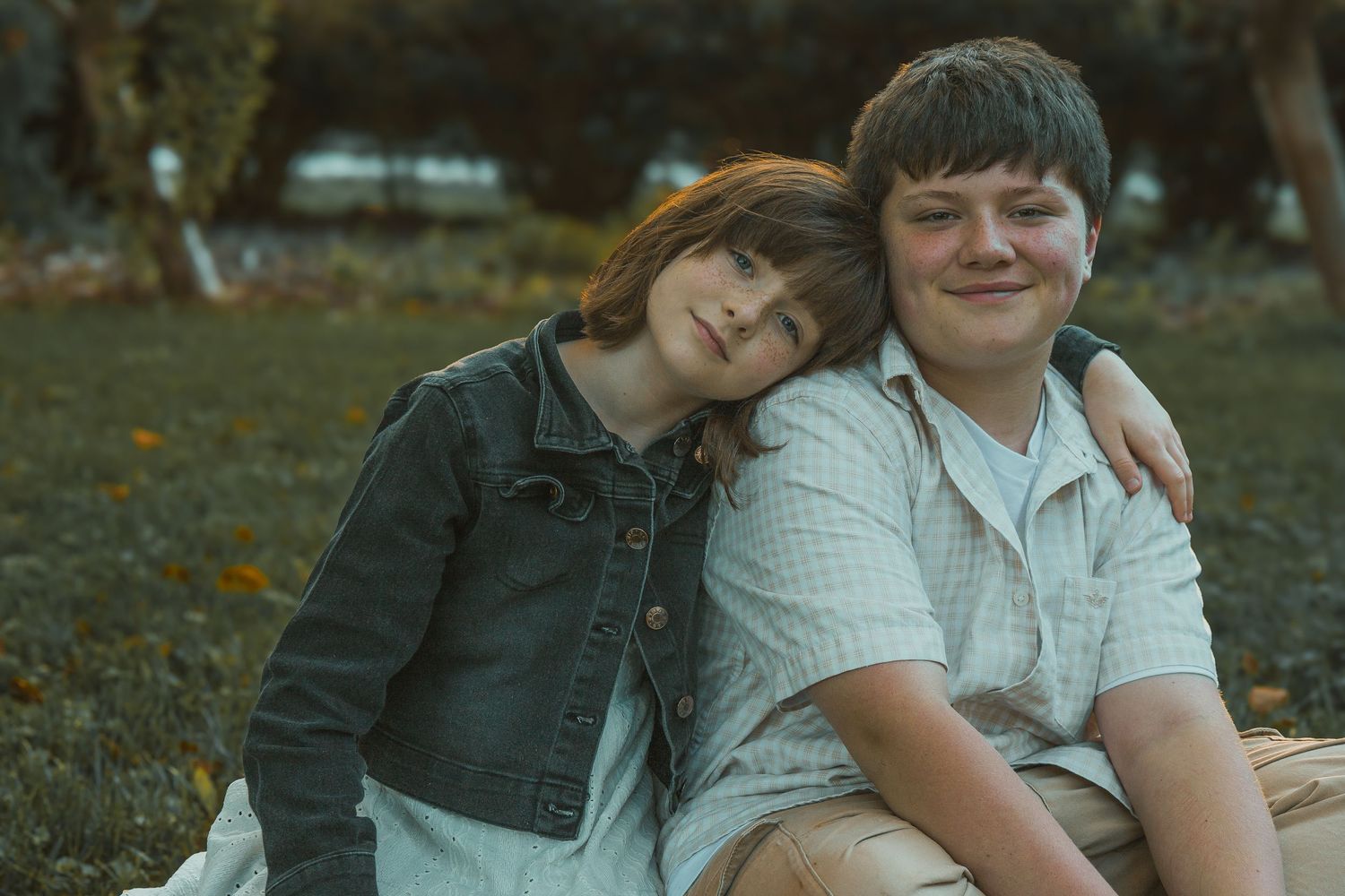 Two siblings in casual clothing sit close together on autumn grass while smiling at the camera.