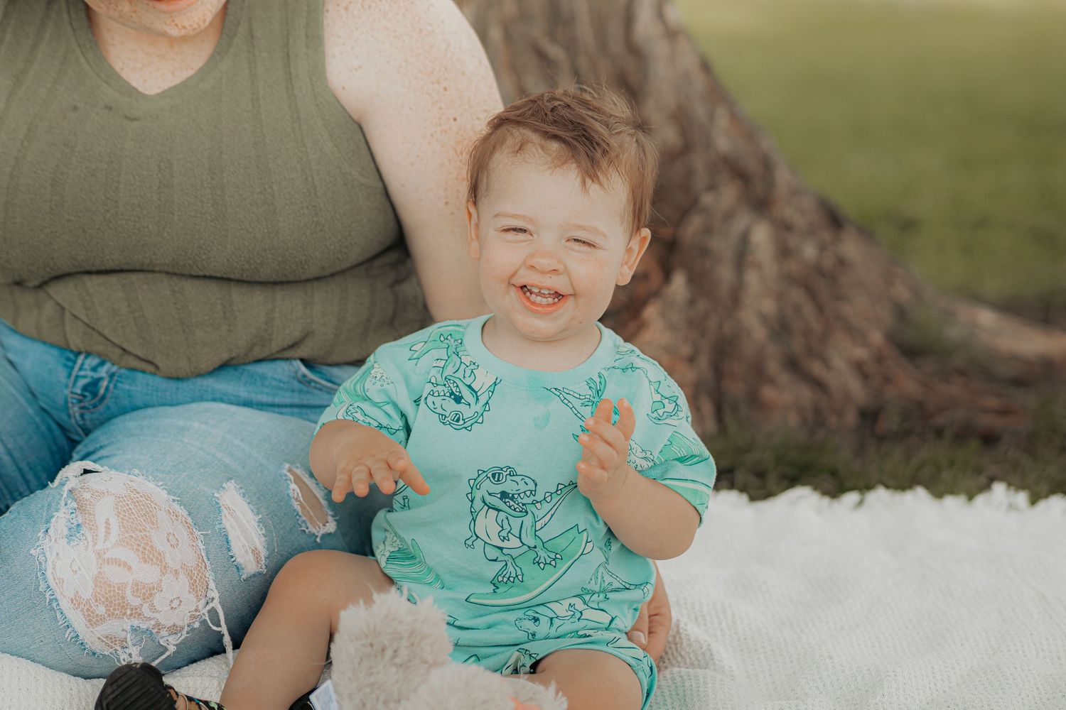 Happy toddler in mint green romper sitting outdoors on white blanket beside a tree, smiling joyfully.