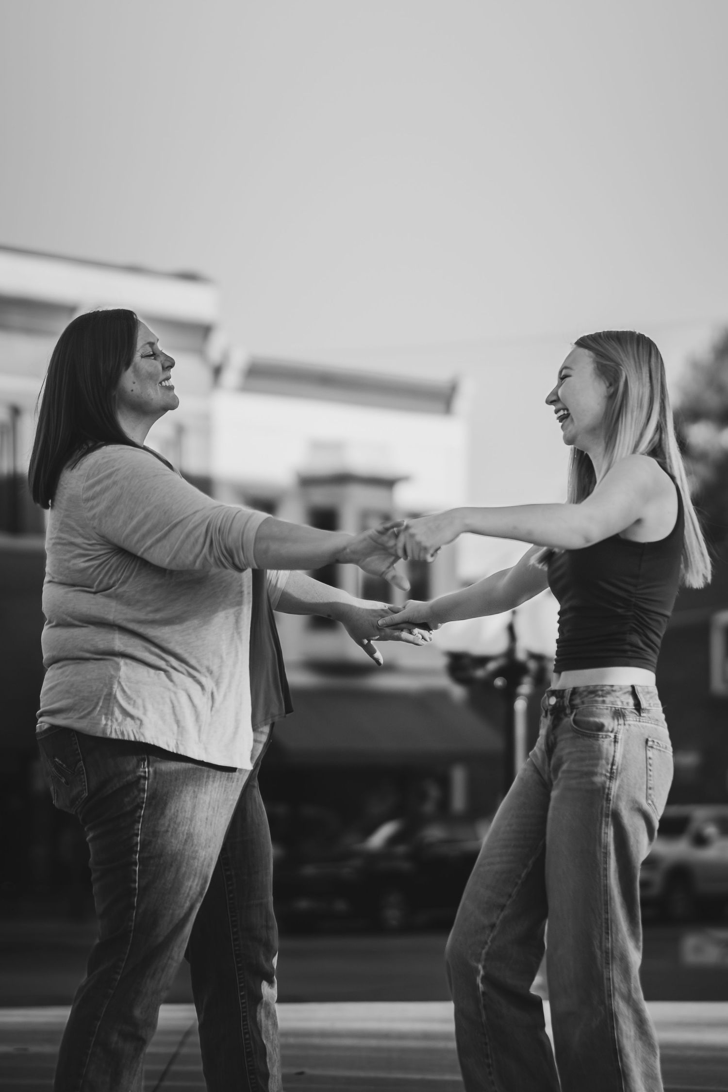 Two women laughing and holding hands outdoors in a black and white photo, sharing a joyful moment together.