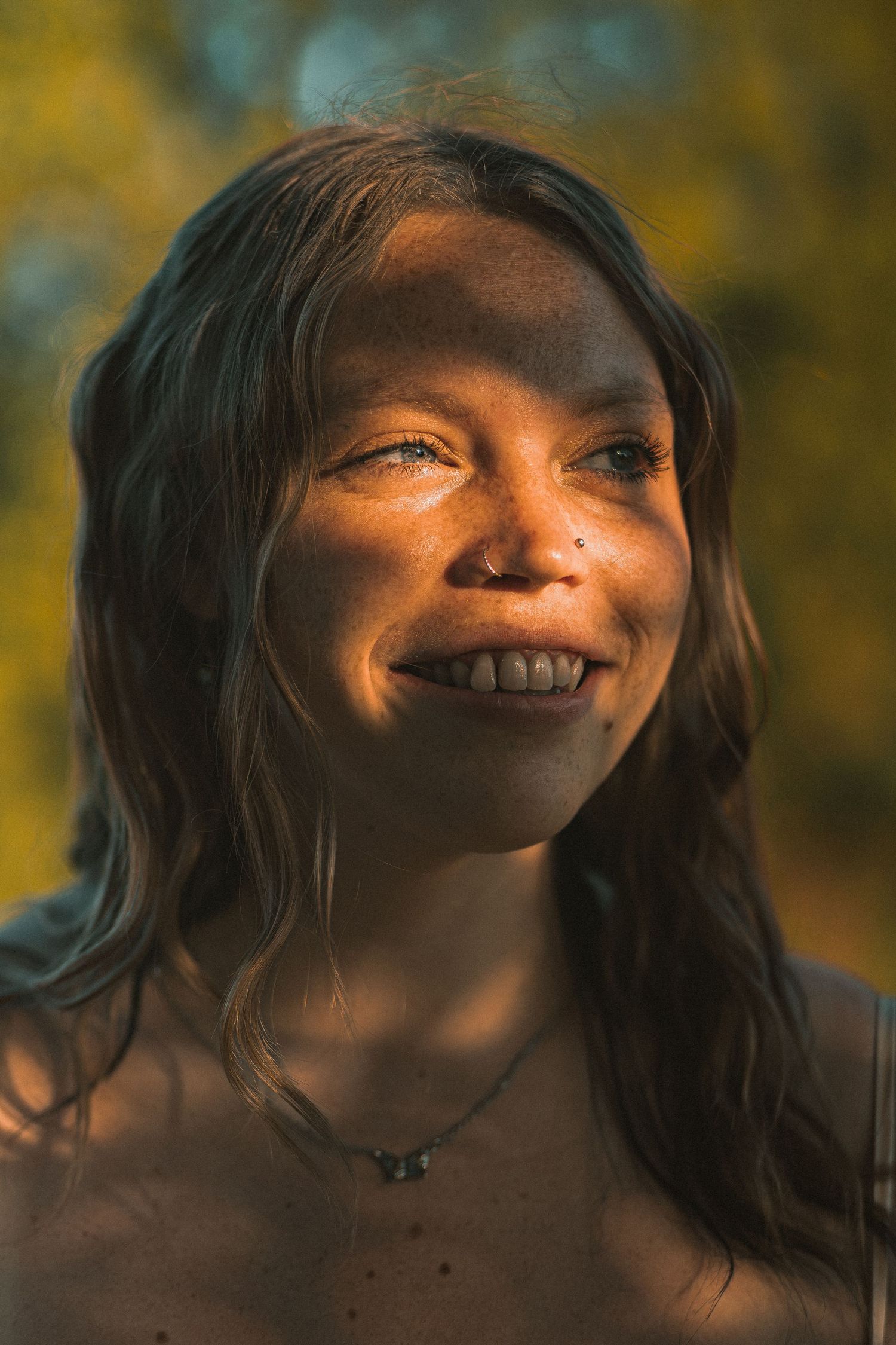Smiling young woman with nose piercing and necklace in warm golden sunlight with blurred green foliage background.