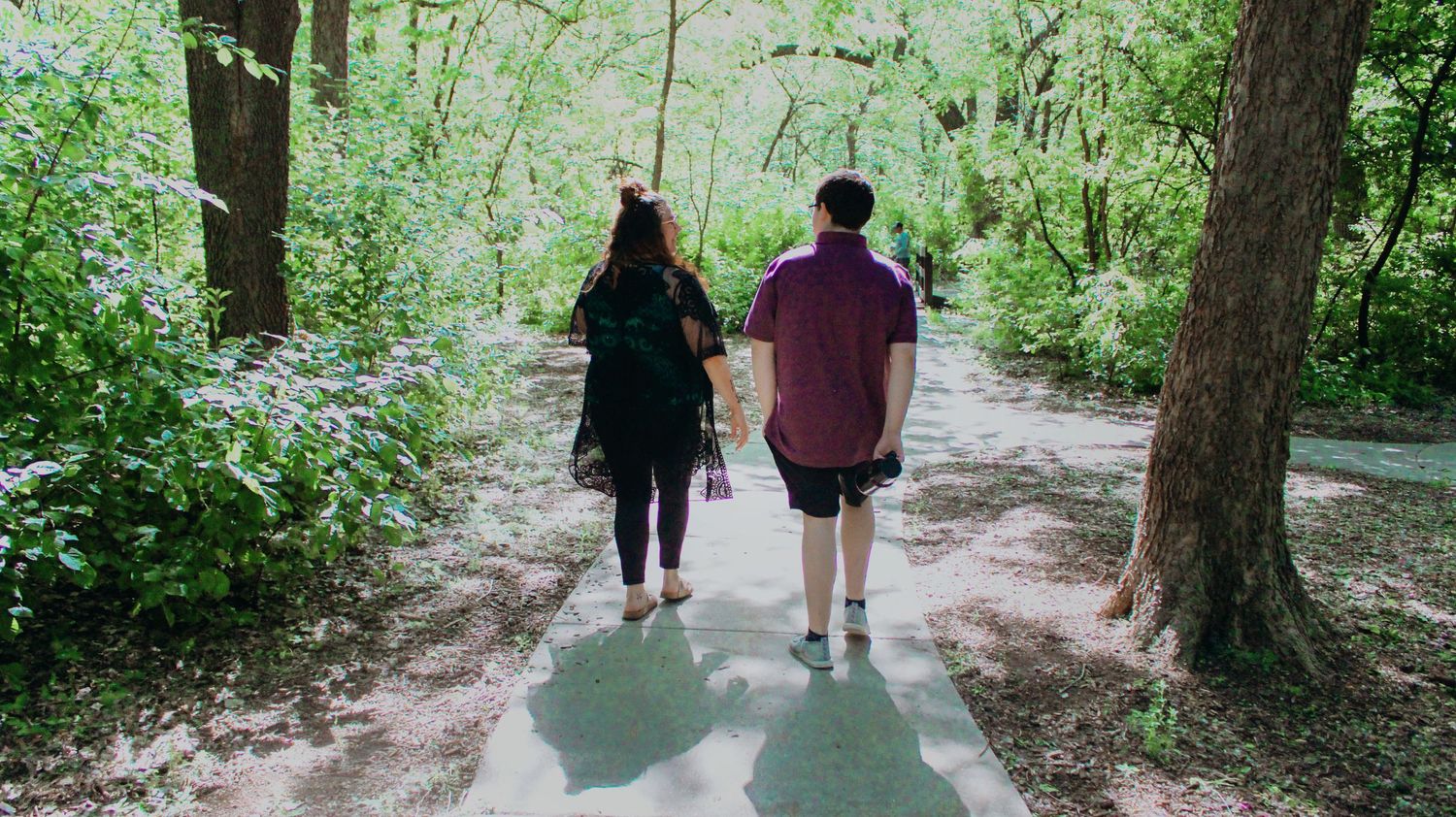 Two people walking together on a shaded forest path surrounded by lush green trees on a sunny day.