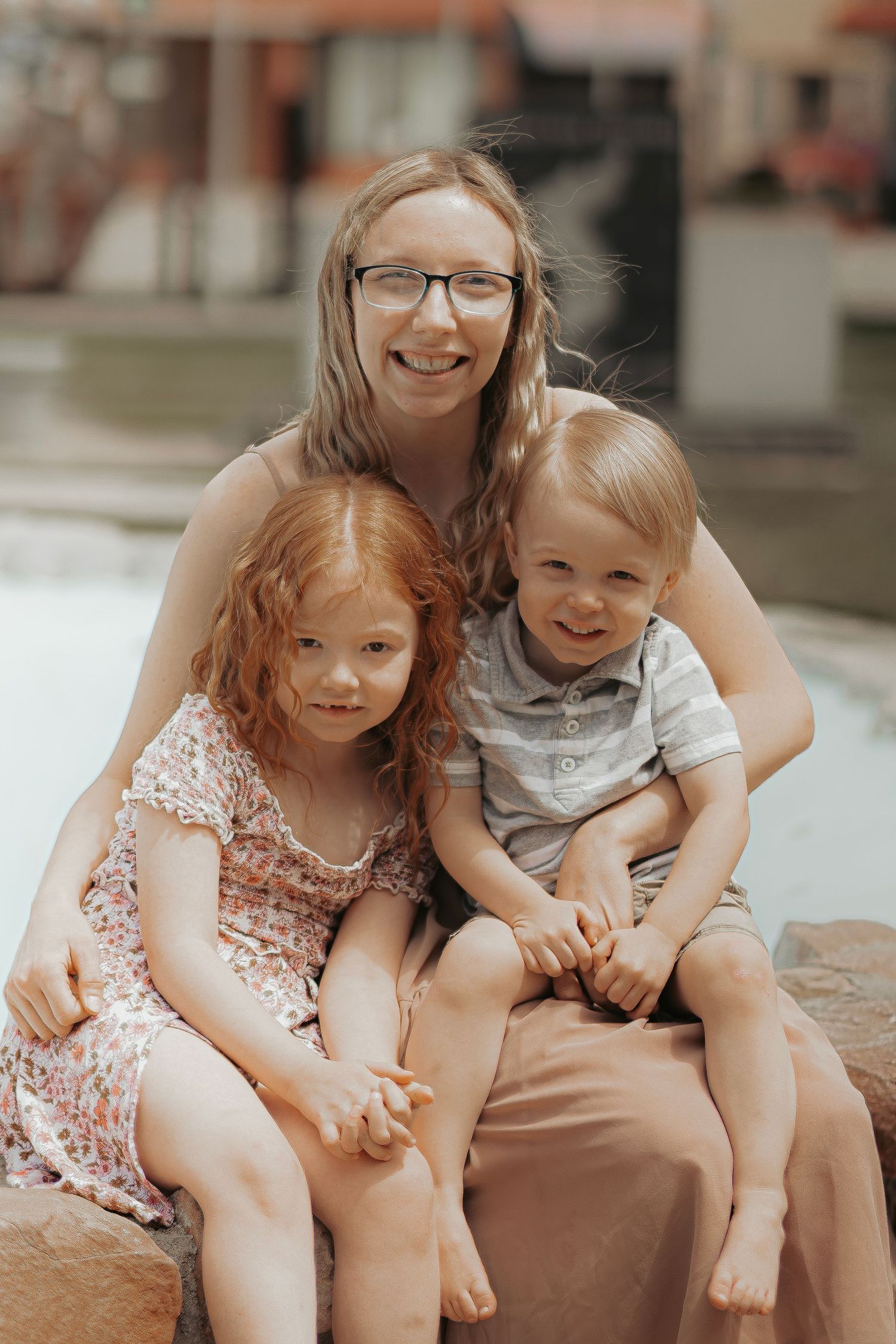 A smiling woman with glasses poses outdoors with two young children, a redheaded girl and a blond boy, sitting together.