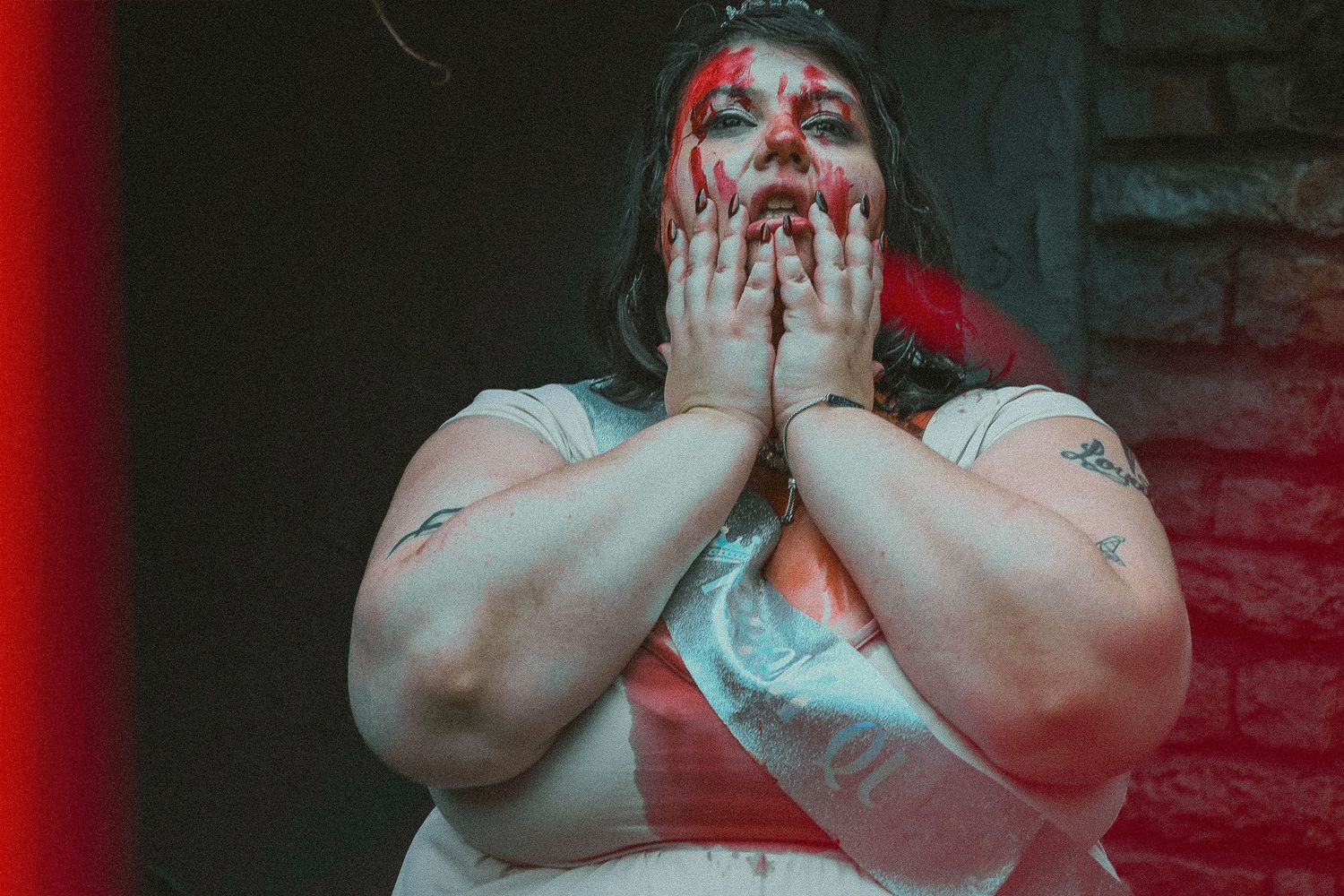 A dramatic portrait lit in red shows someone in white clothing with theatrical horror-style makeup covering their face.